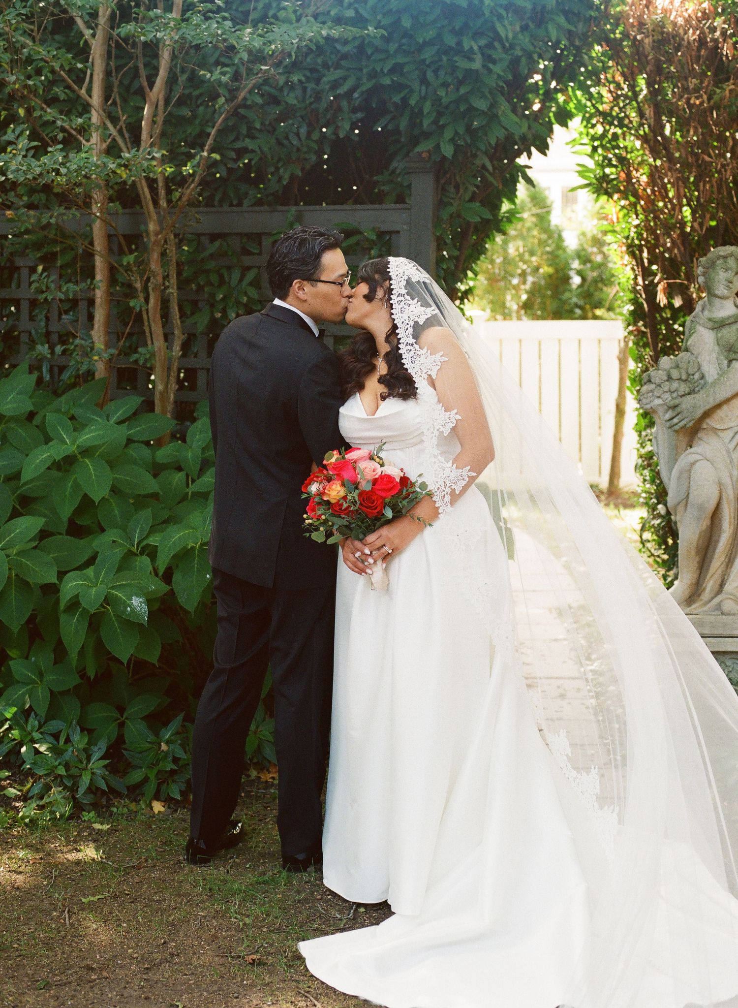 A wedding couple poses in a garden setting with greenery and white flowers during an outdoor ceremony.