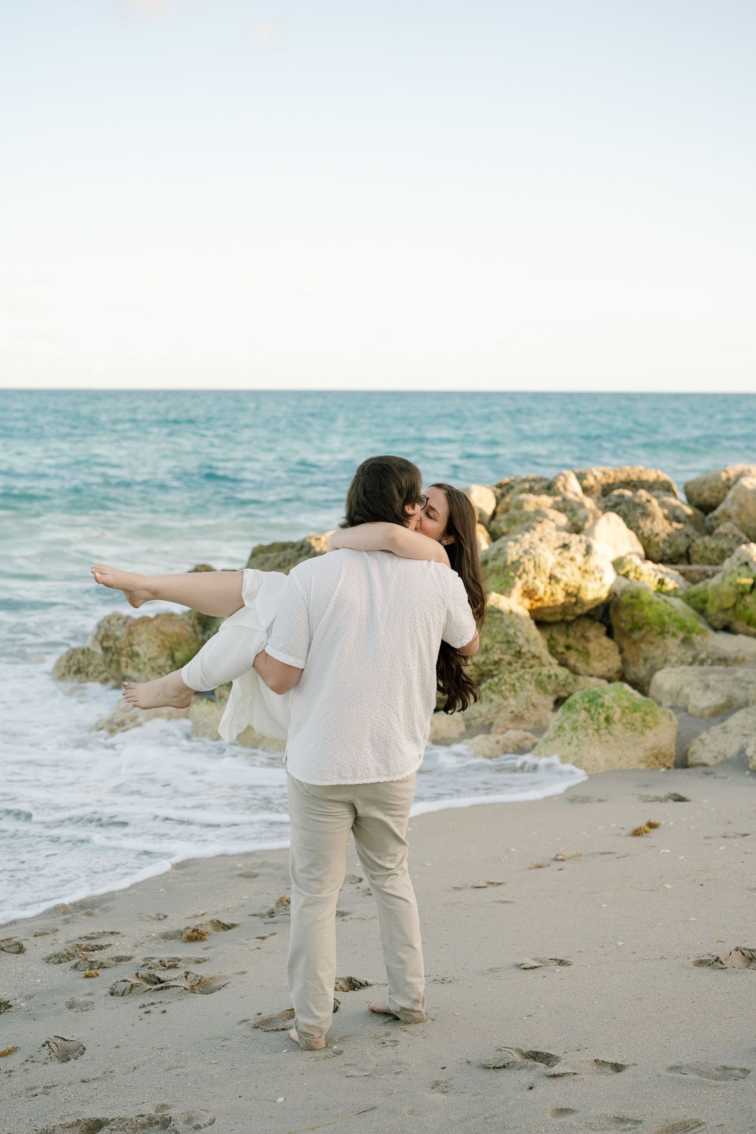 A couple in white outfits dances playfully on a beach with rocky outcrops and ocean waves in the background.