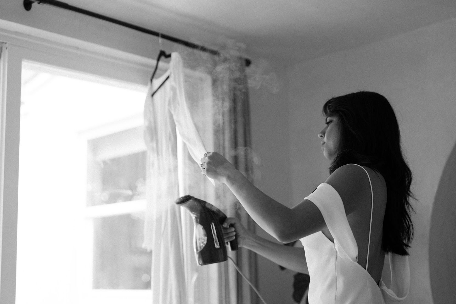 Black and white photo showing silhouettes preparing in a bright room with curtains.