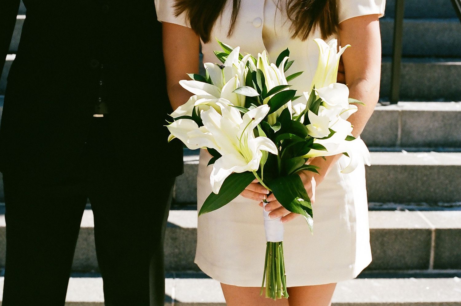 A bride holds a white lily bouquet while wearing a short white dress and standing in front of a window.