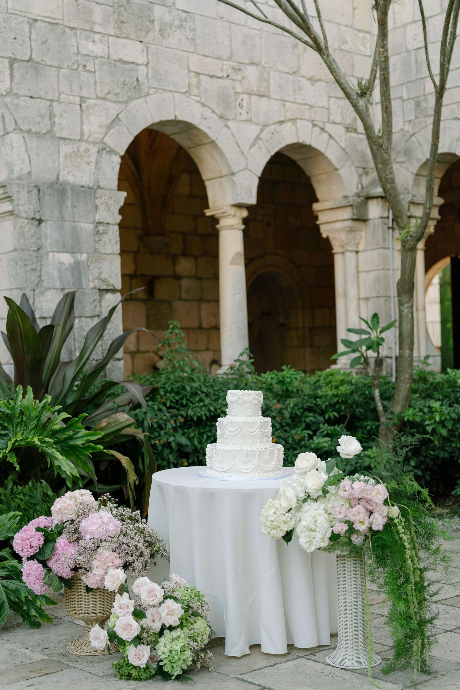 Elegant four-tier white wedding cake displayed on draped table with pink floral arrangements in stone courtyard.