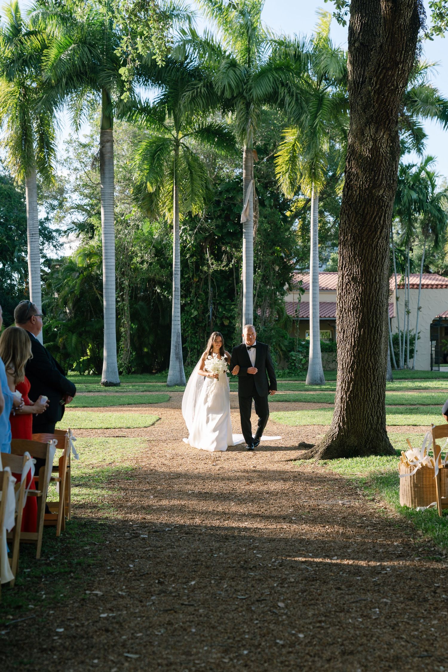 Bride in white ball gown walks down tree-lined aisle with escort at outdoor garden wedding ceremony.