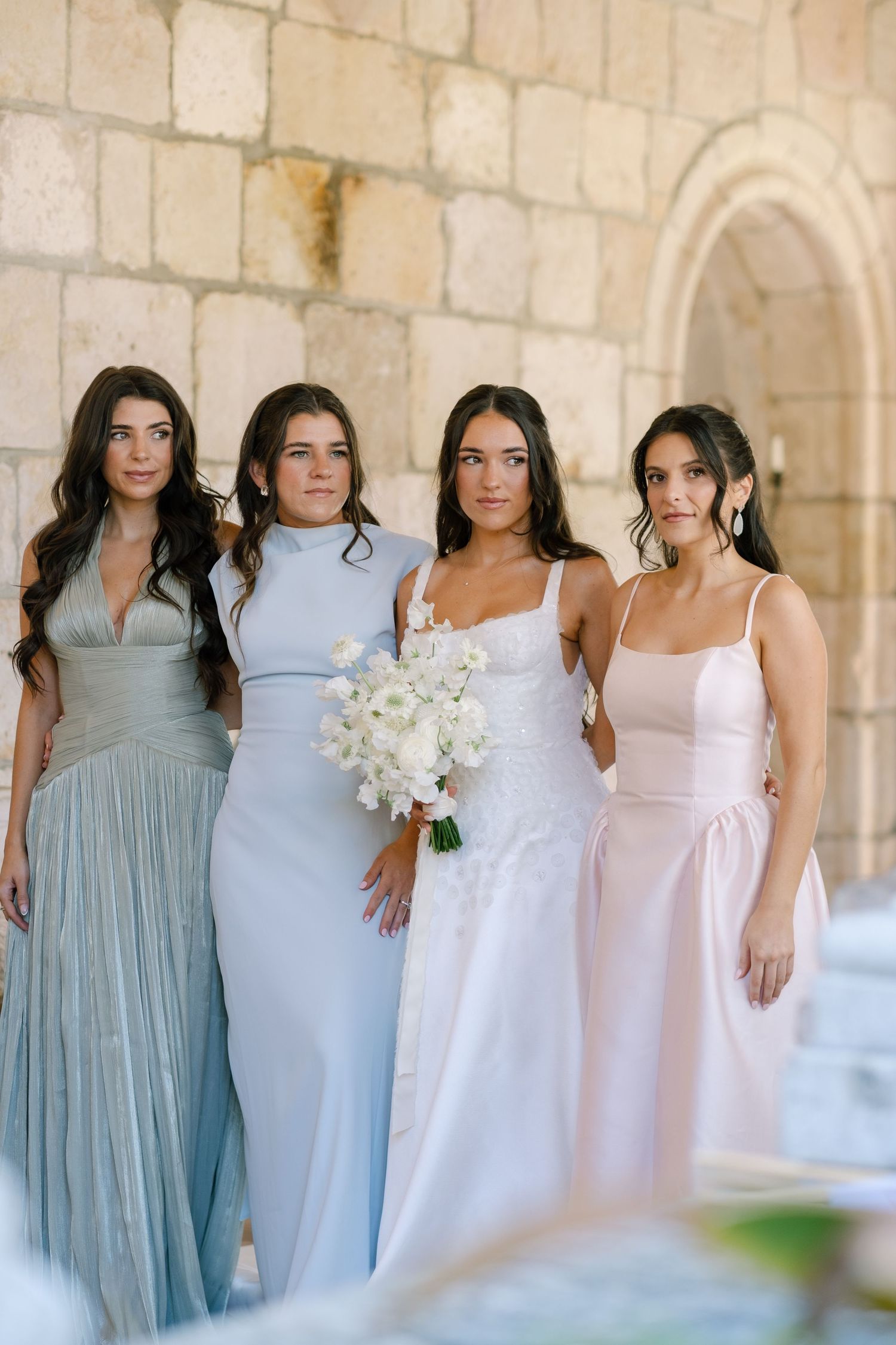 Bride holding white bouquet poses with three bridesmaids in pastel gowns before stone archway.