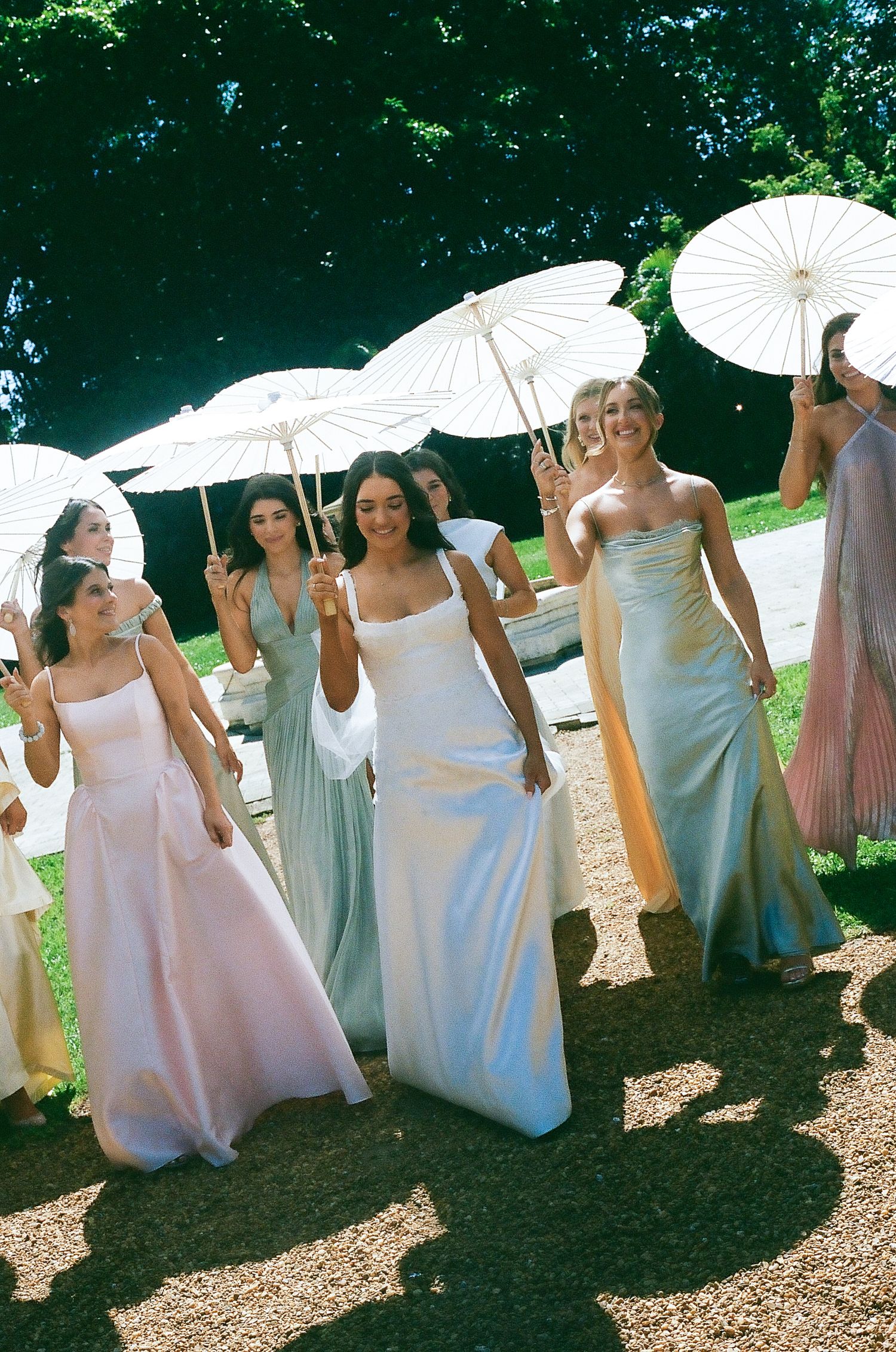 Bride in white gown walks with bridesmaids in pastel dresses holding white parasols outdoors.