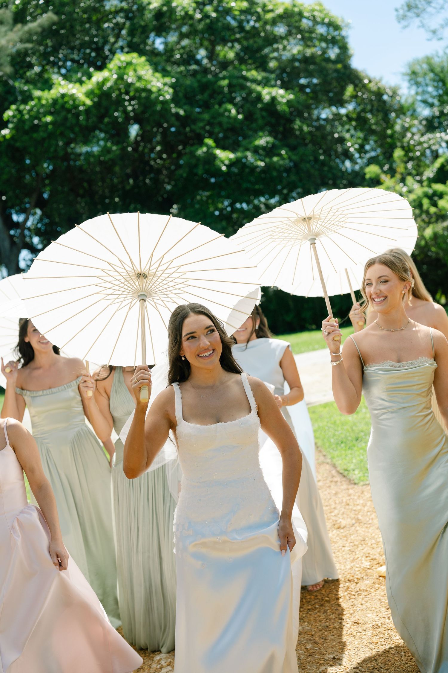 Bride and bridesmaids in sage green gowns walking outdoors holding white parasols on sunny day.
