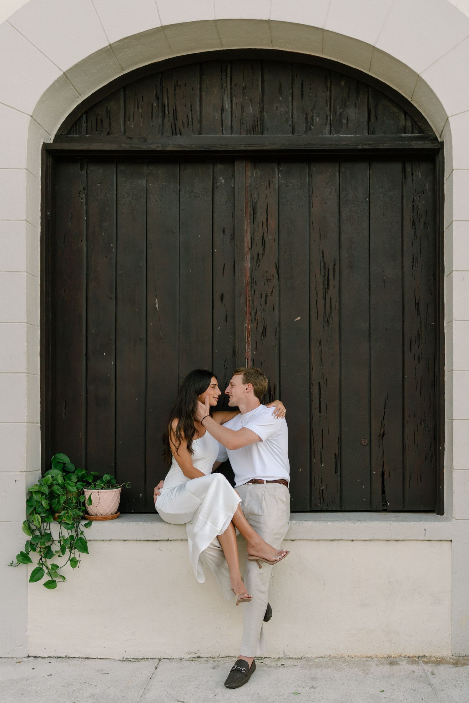 Romantic couple embracing on white ledge before dark arched wooden door, woman in white dress sitting on mans lap.