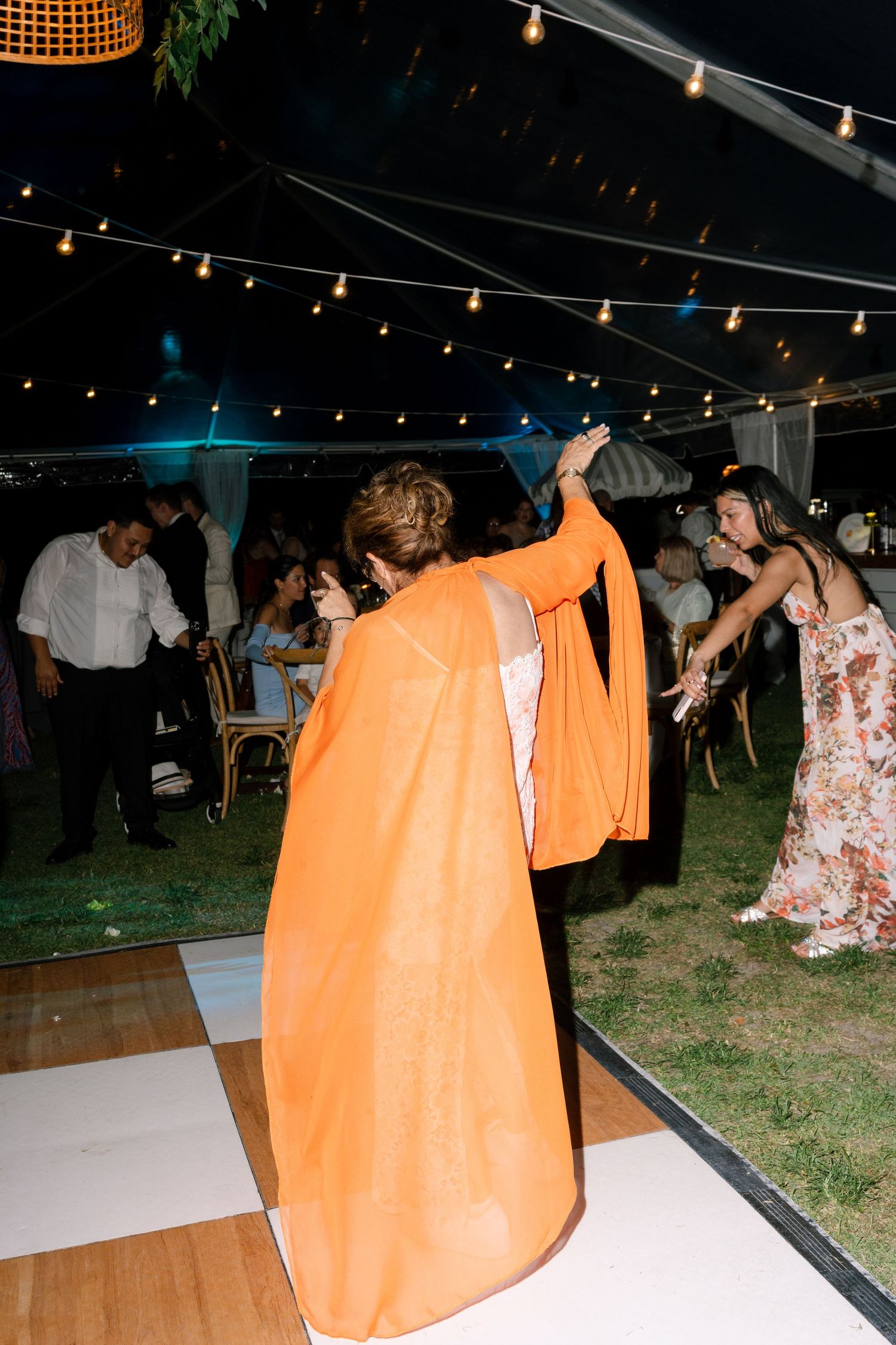 Woman in orange cape dress dancing at an outdoor wedding reception under string lights at night.