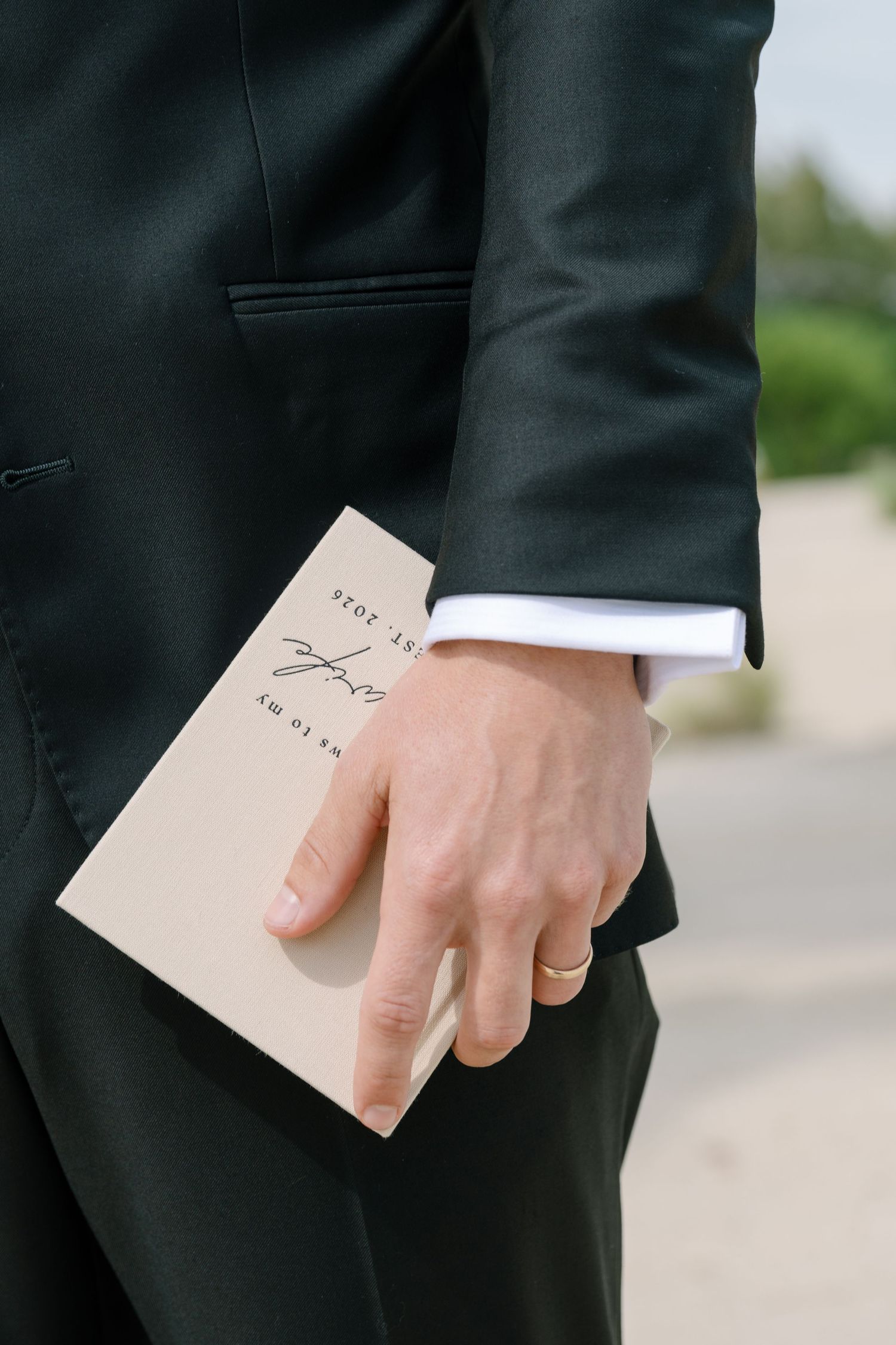 Groom in black suit holding wedding vows booklet with gold wedding ring visible on hand.