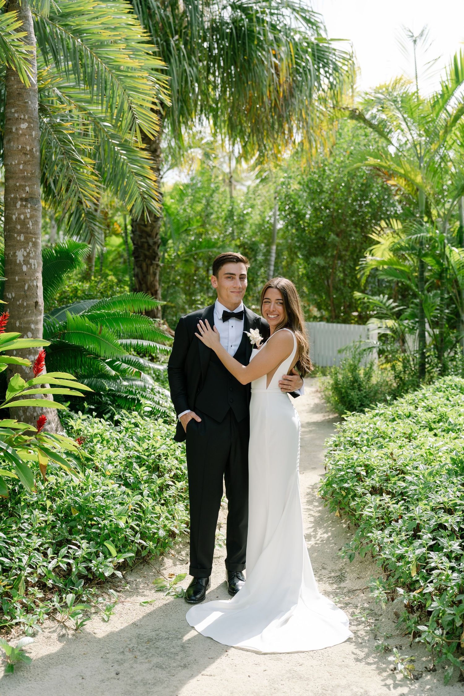 Bride and groom posing together on tropical garden path, groom in black tuxedo, bride in sleek white gown.
