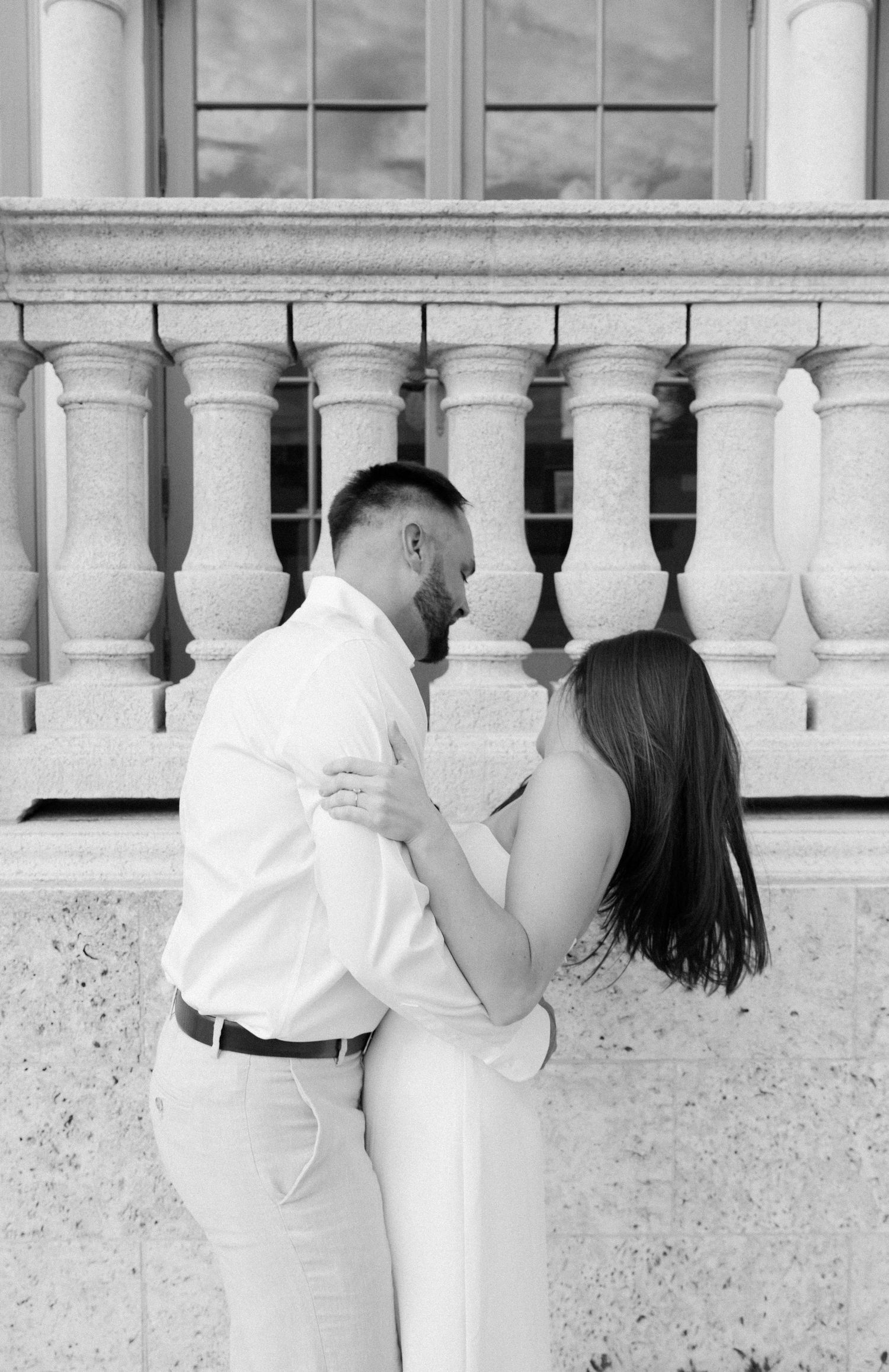 Black and white photo of a couple in white outfits embracing by an ornate stone railing with columns.