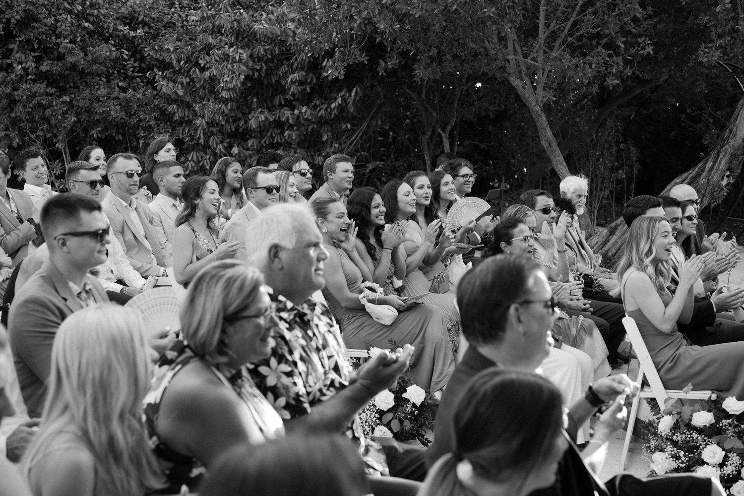 Black and white photo of a crowd sitting outdoors watching an event, with people attentively looking off to one side.