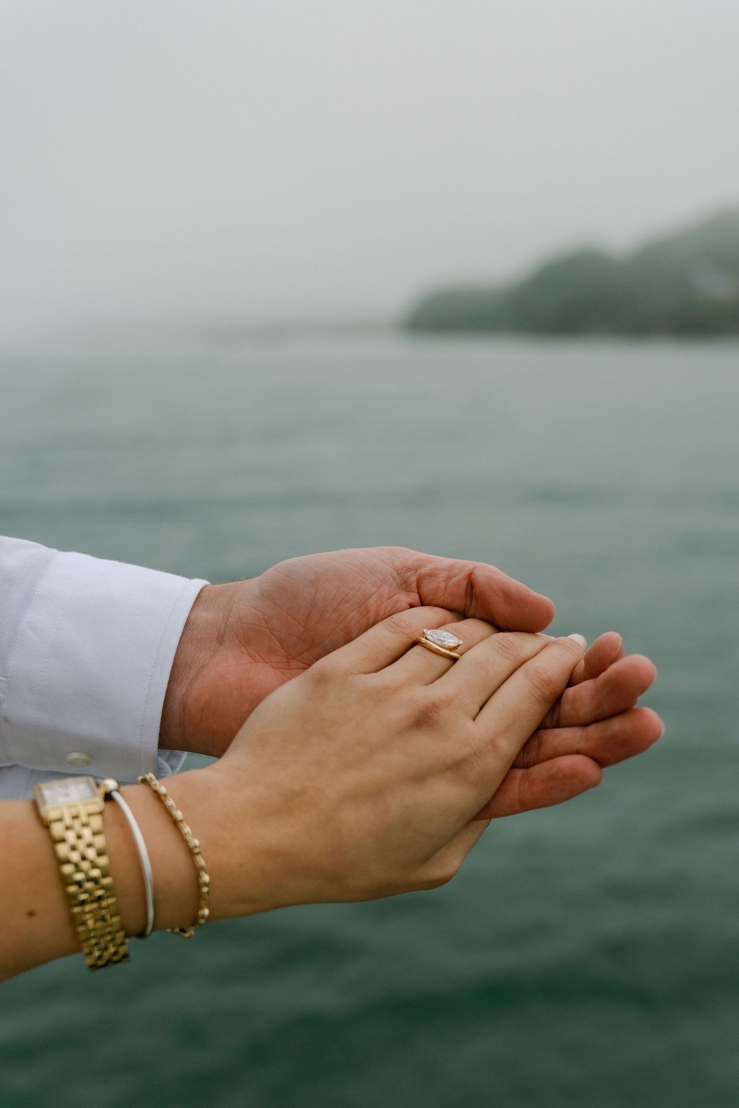 Two hands gently clasped together near water, showcasing a diamond engagement ring and gold jewelry.