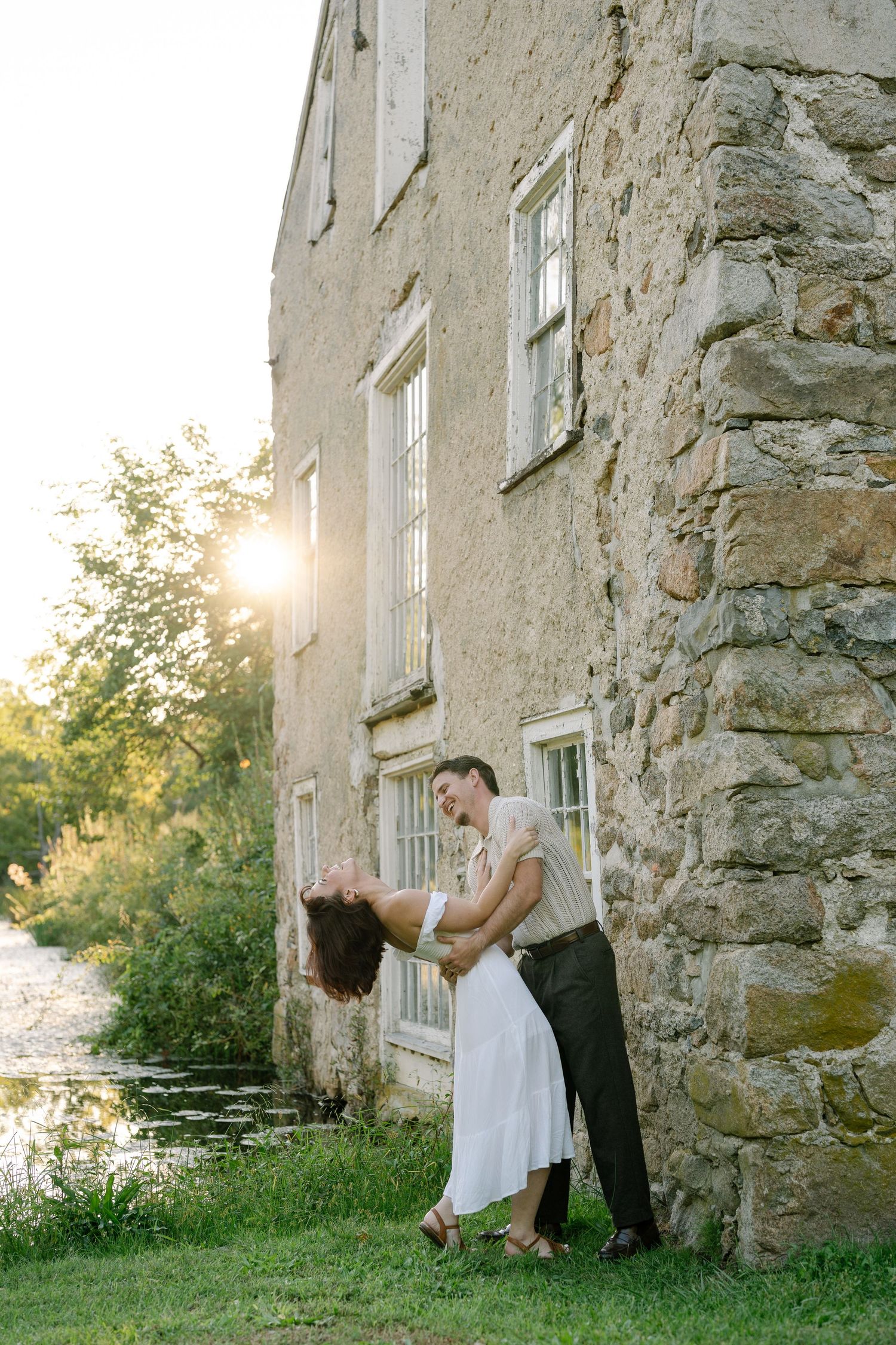 Couple dancing romantically beside a rustic stone building at golden hour sunset with lush greenery.