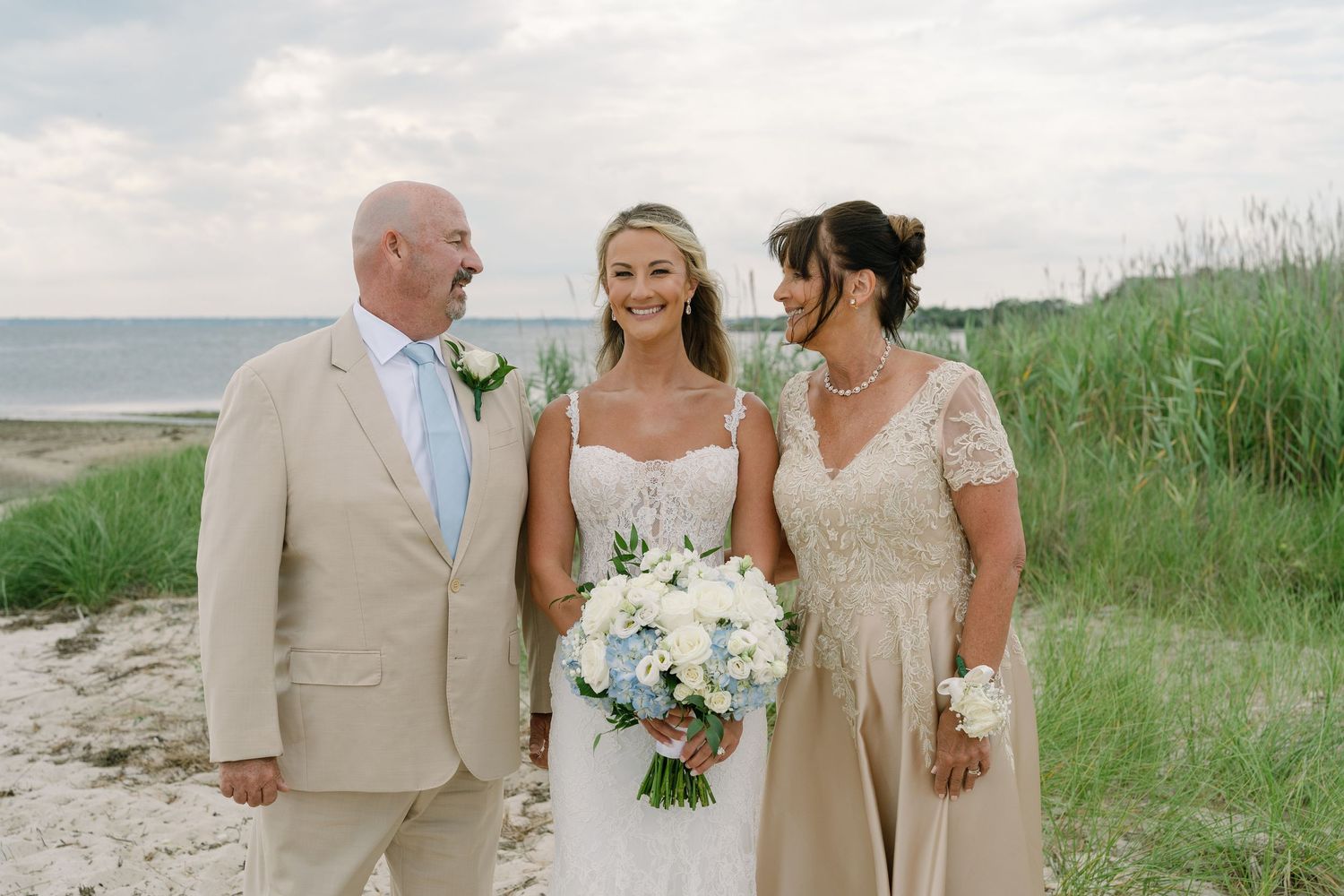 Bride in white gown holds bouquet flanked by parents on sandy beach with ocean backdrop.