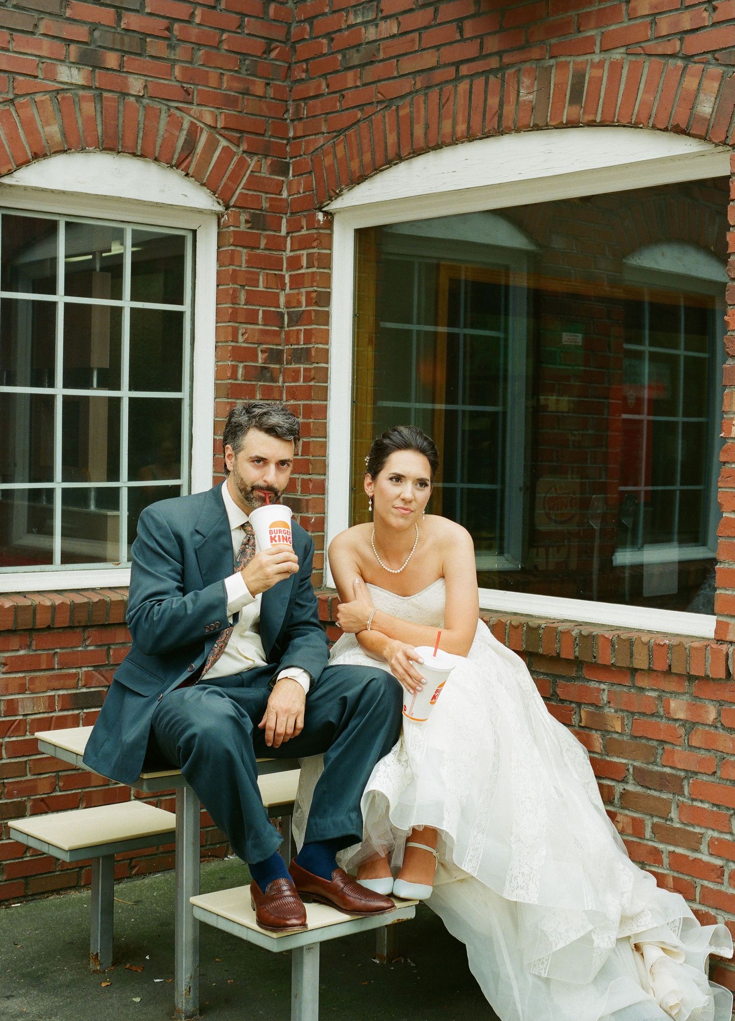 Wedding couple shares candid moments while sitting on a bench by a brick building.