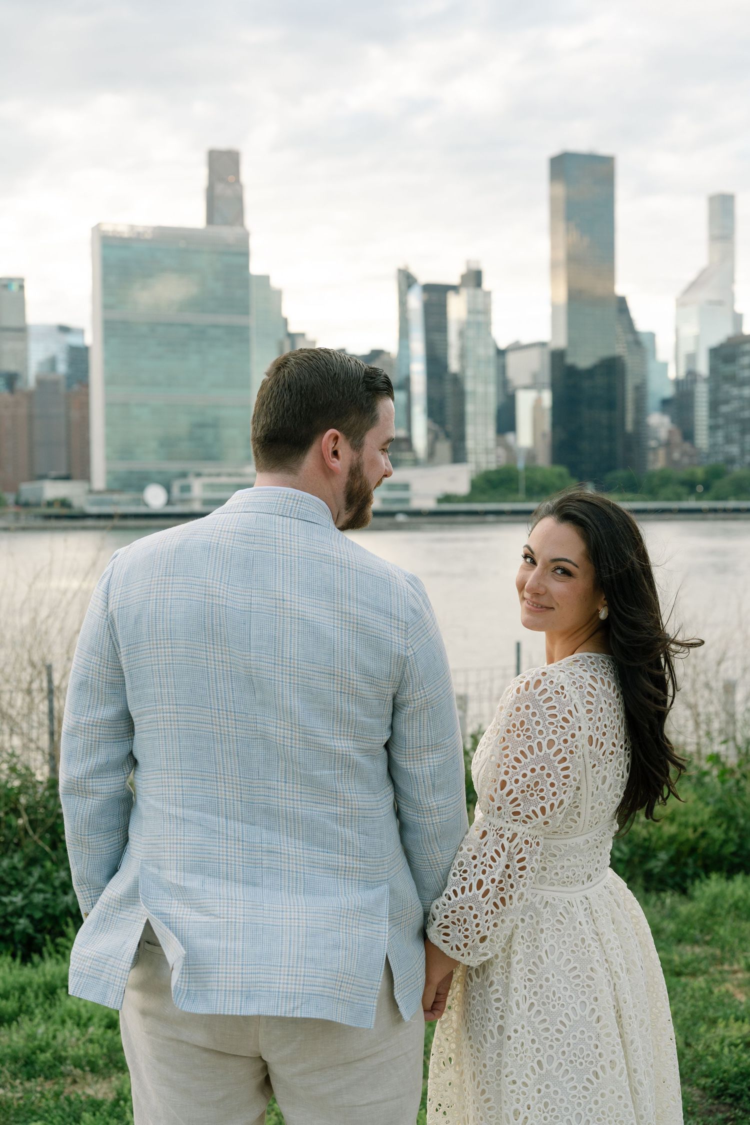 Couple holding hands with NYC skyline backdrop, woman in white lace dress smiling over shoulder.