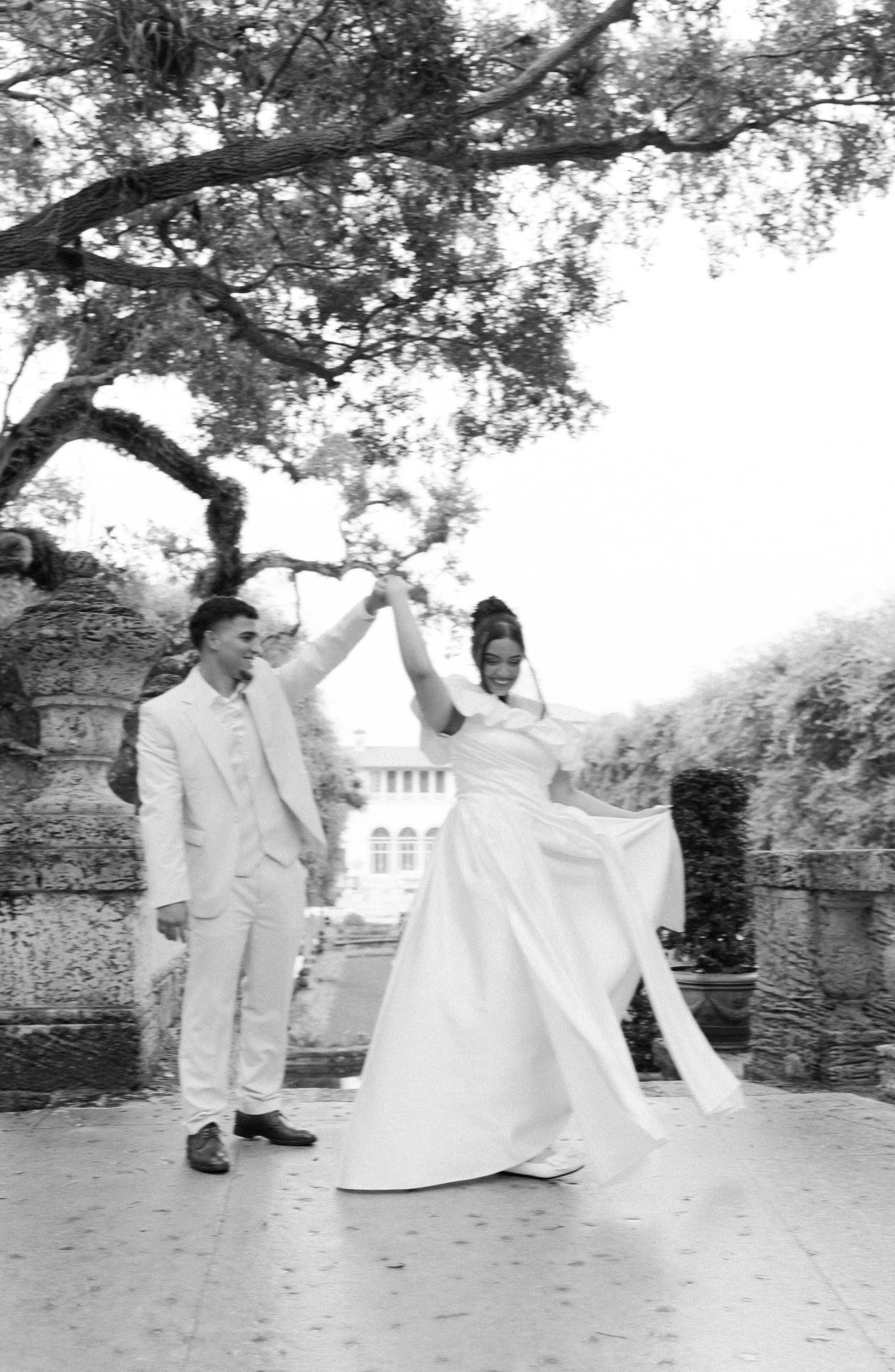 A couple spins with joy in a black and white wedding photo, set against a stone fence and lush trees.