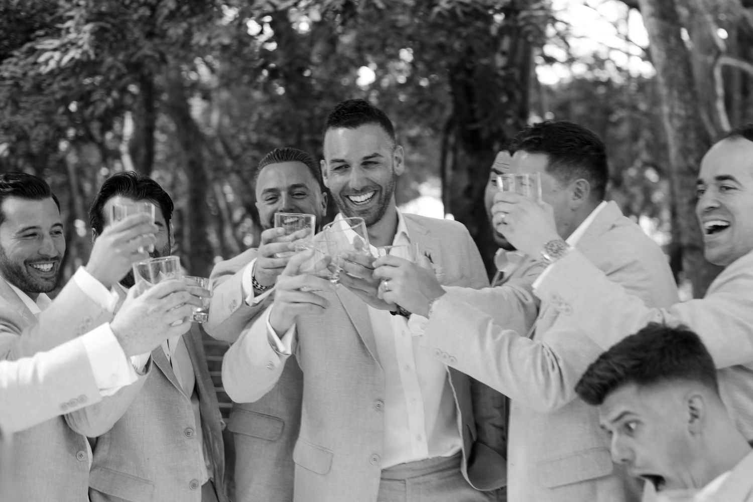 A group in light colored suits toasts with drinks at an outdoor celebration in black and white photography.