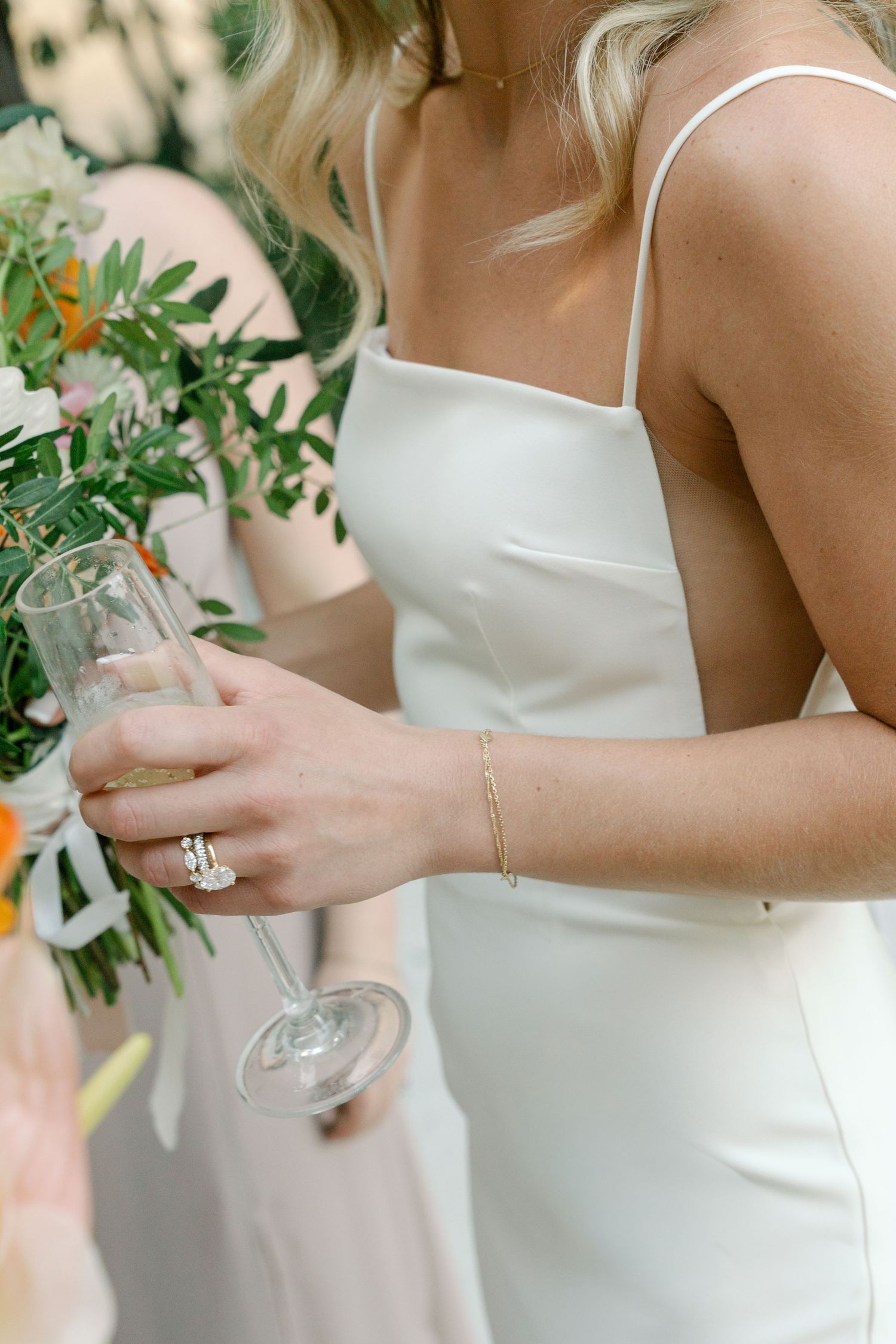 Wedding party members in white dresses holding champagne glasses with greenery in background.