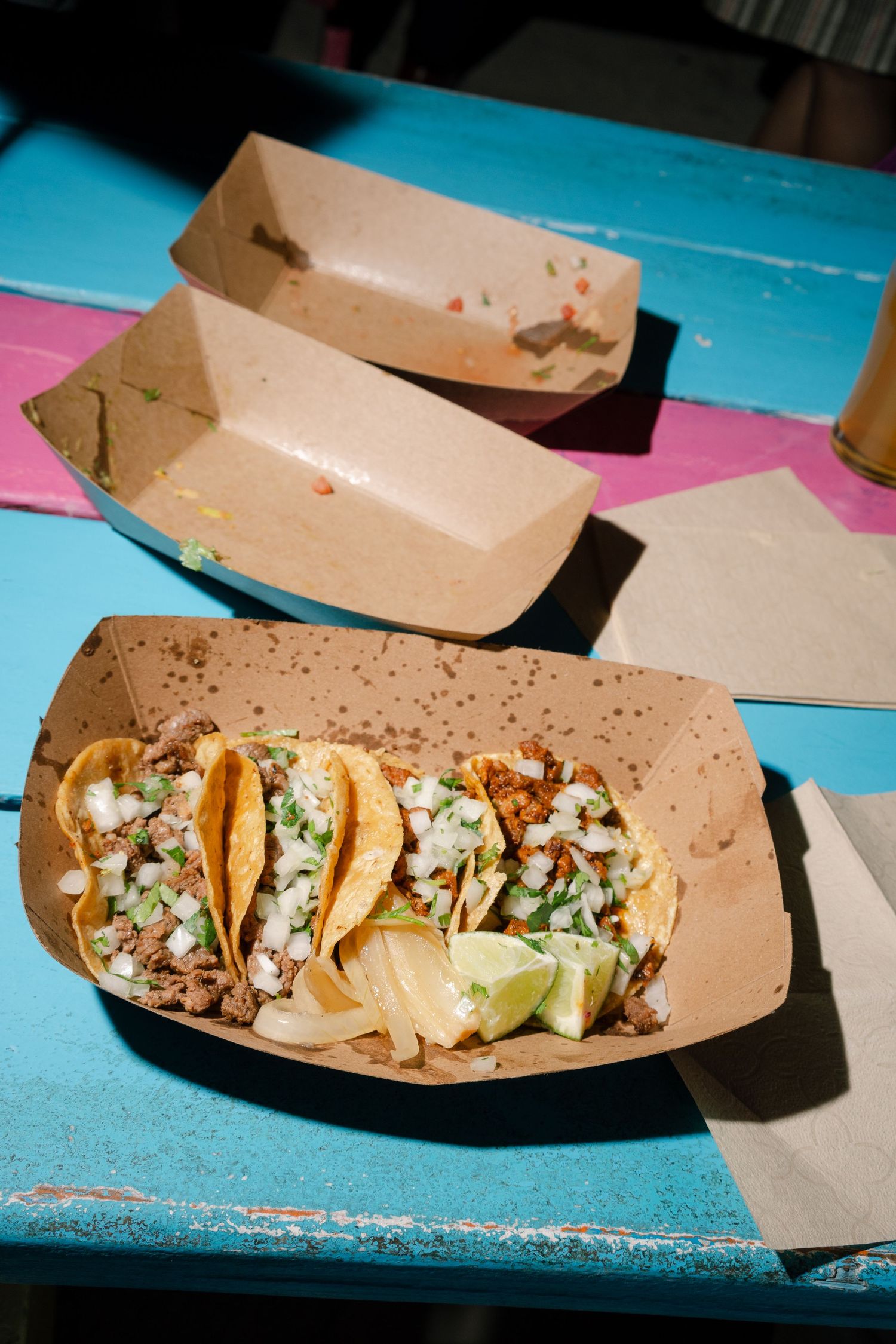 Street tacos served in brown paper boats on a bright blue table.