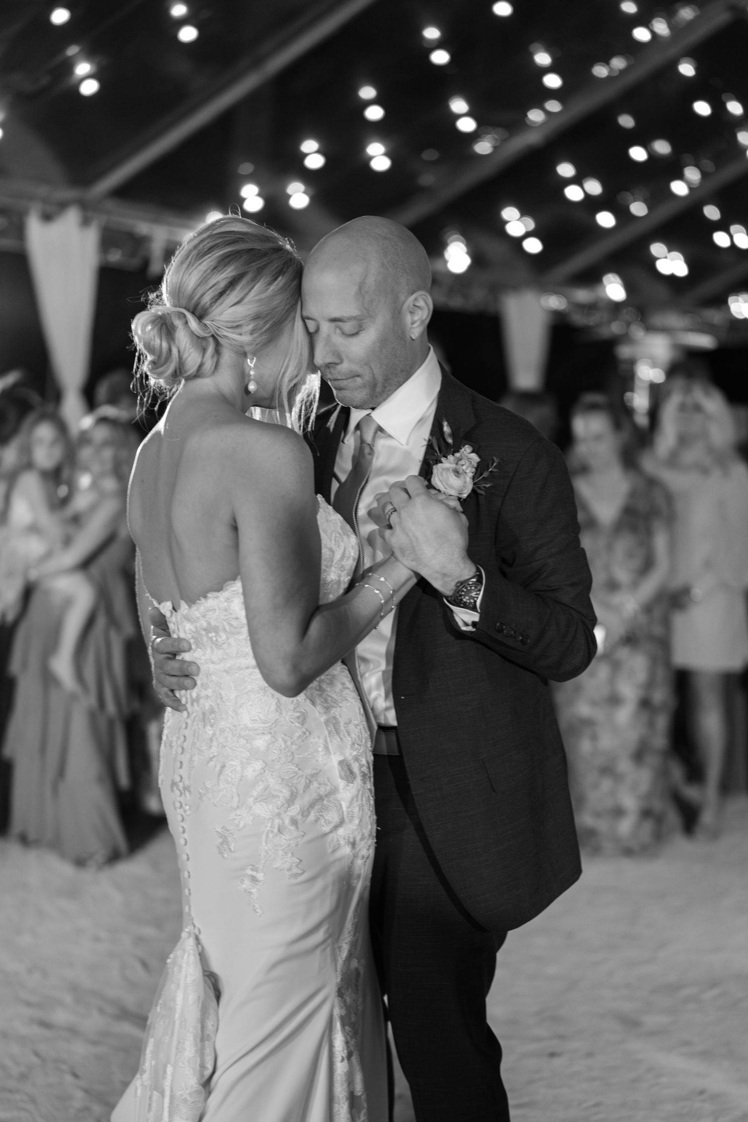 A couple shares their first dance at a beach wedding reception under twinkling string lights at dusk.