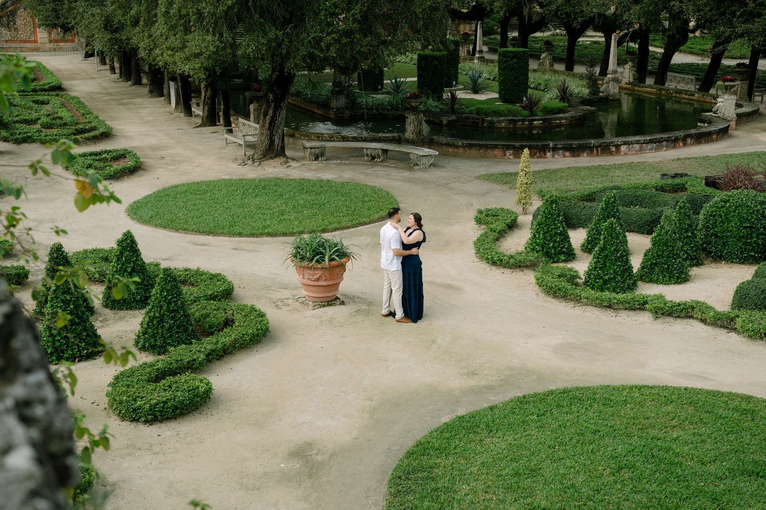 Manicured garden pathway with ornamental hedges and trees leads through a formal landscaped park on a sunny day.