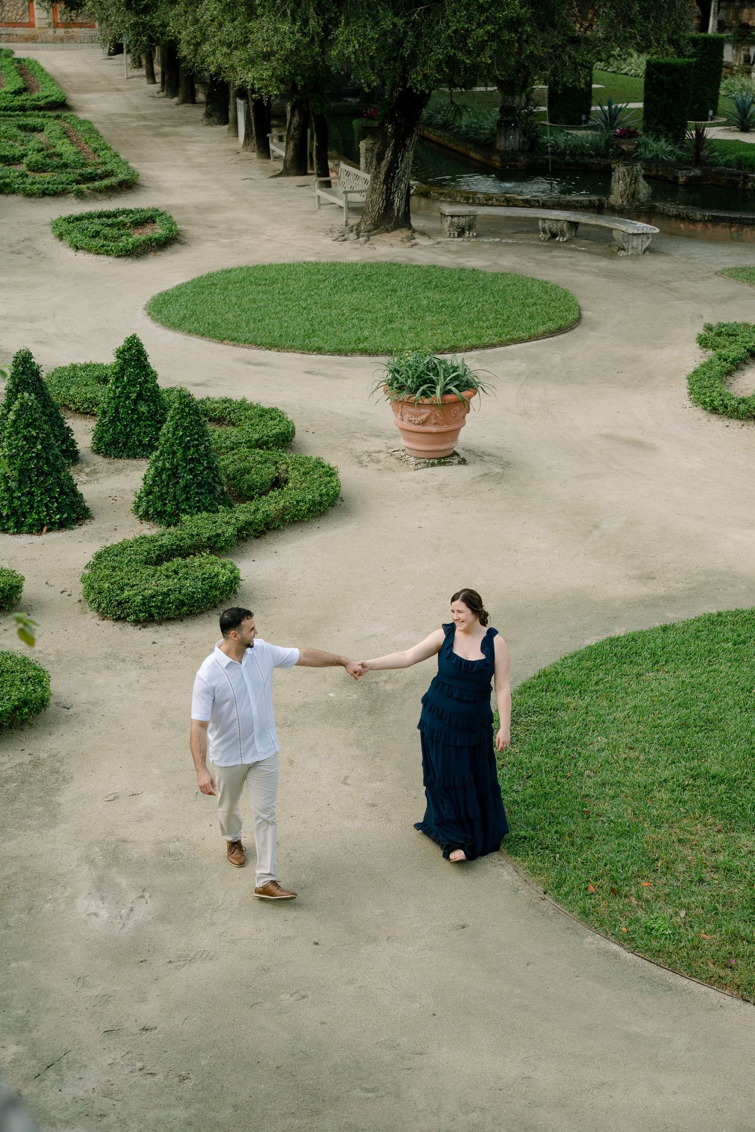 Two people walk along a garden path with manicured hedges and trees in an elegant outdoor setting.