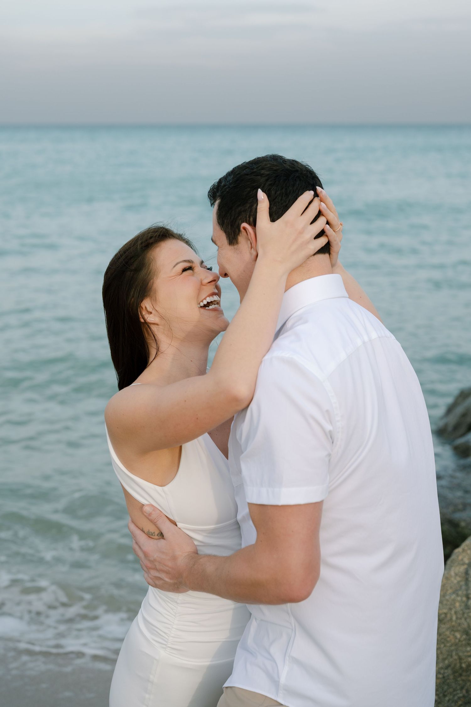 A couple shares a romantic moment by the ocean in matching white outfits.