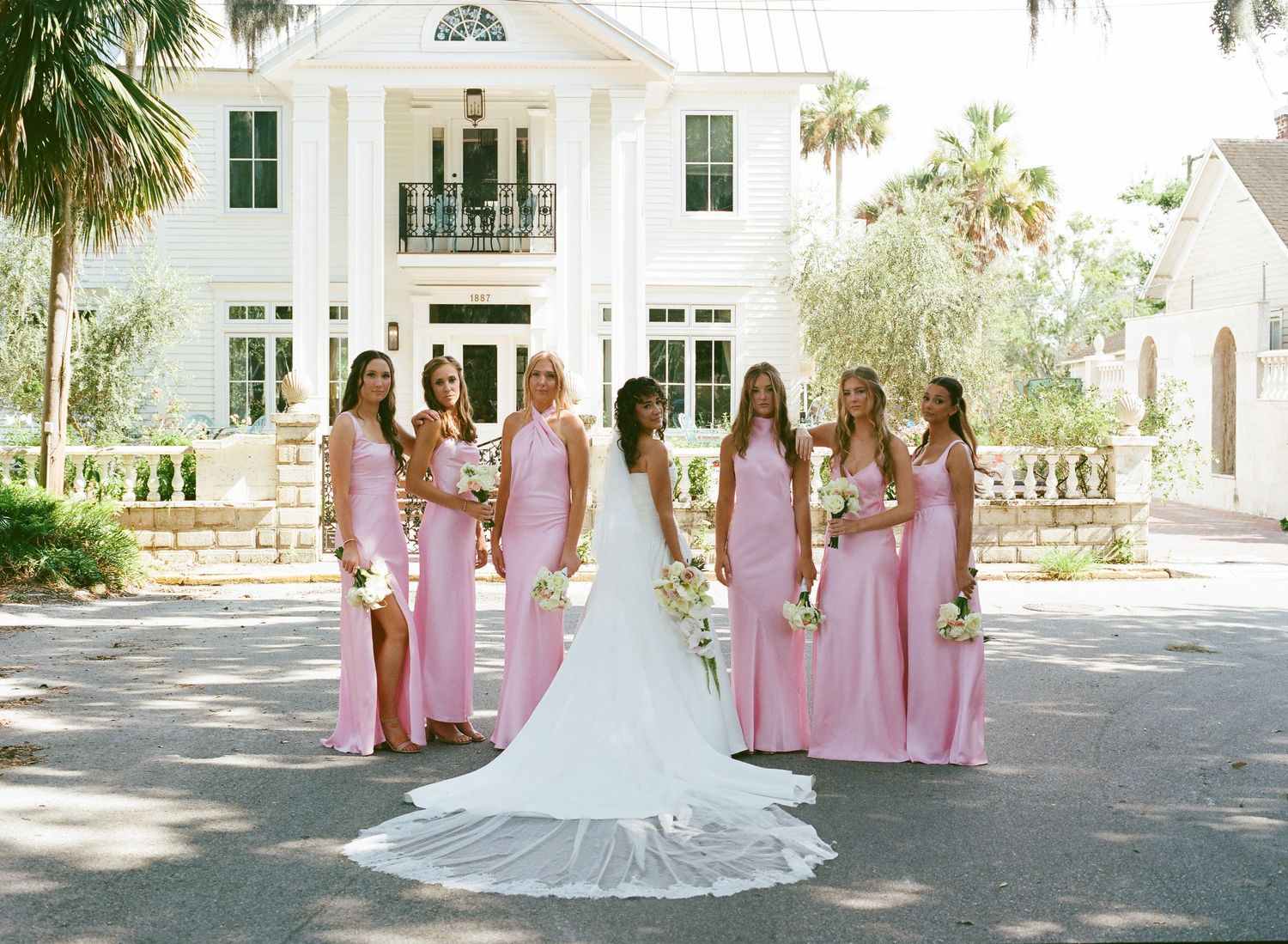 A wedding party in pink dresses poses outdoors at an elegant white mansion venue with palm trees.