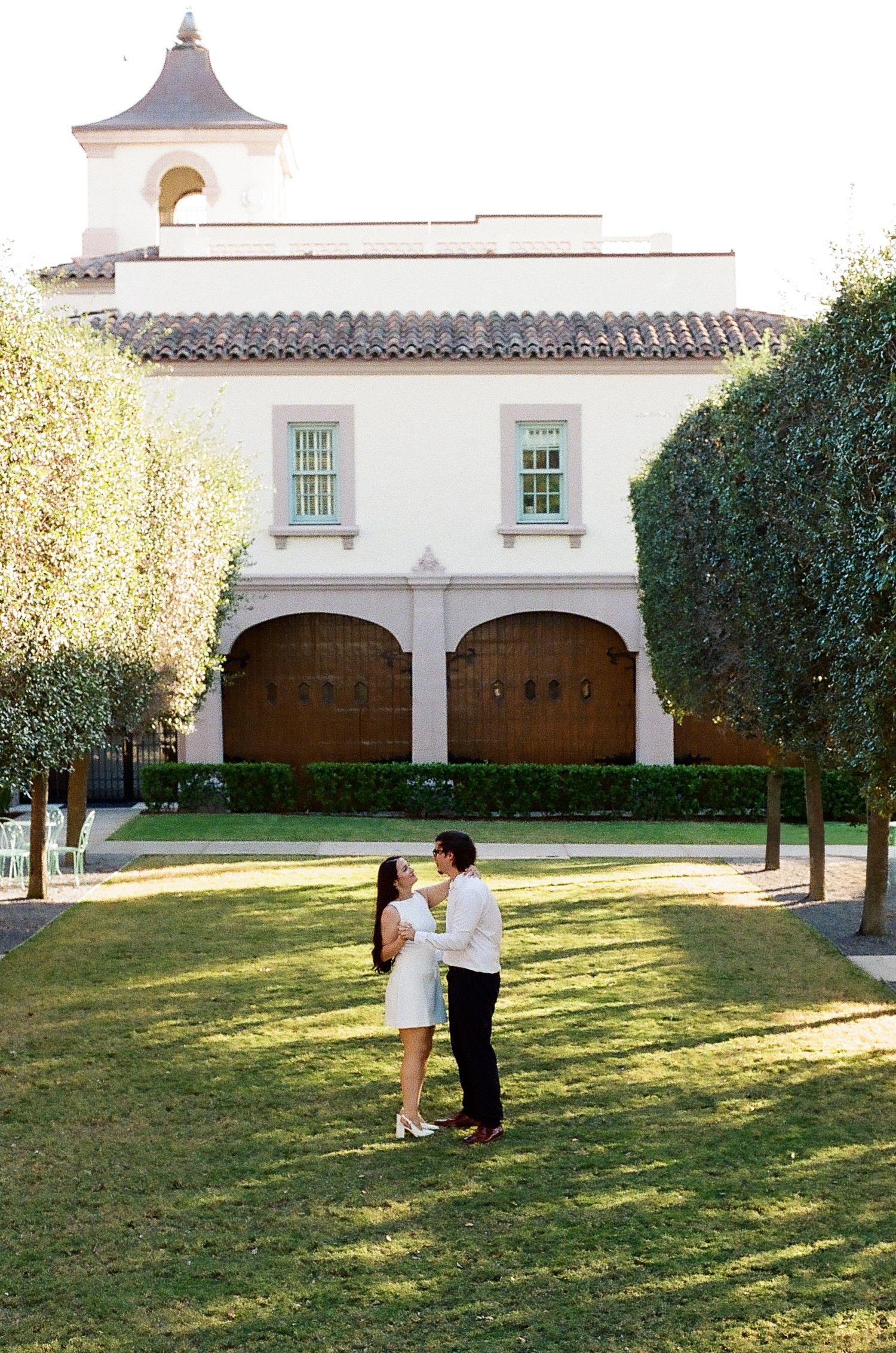 A couple shares a romantic moment on a manicured lawn in front of a Spanish-style building with white arches.