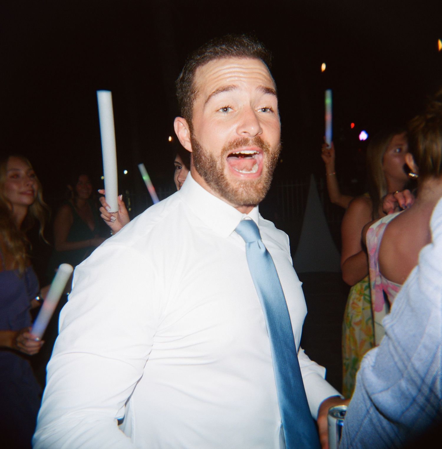Wedding guests dance and celebrate at a lively reception party with glowing lights in the background.