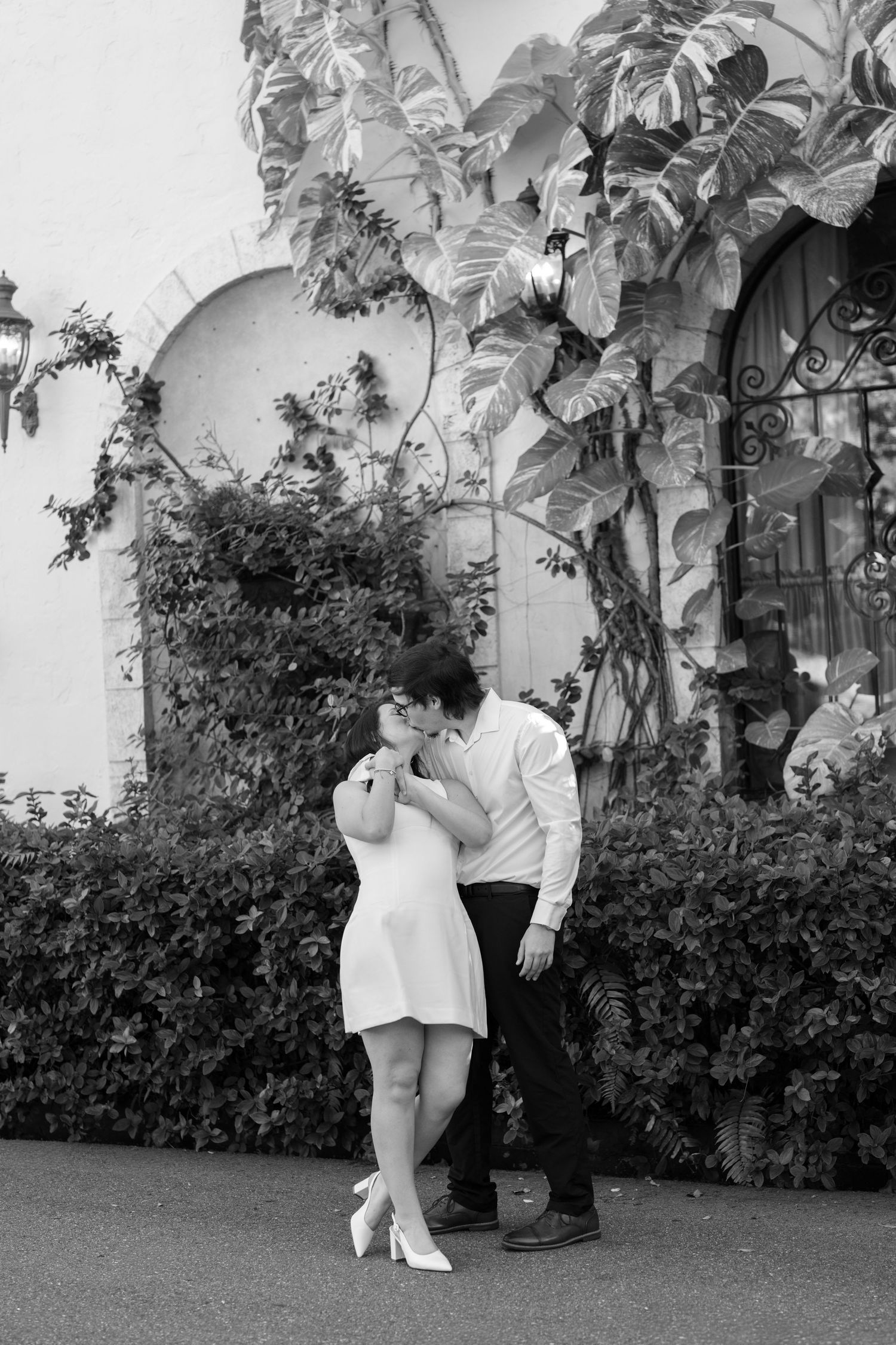 A romantic black and white photo of a couple embracing by a vine-covered wall with an arched doorway.