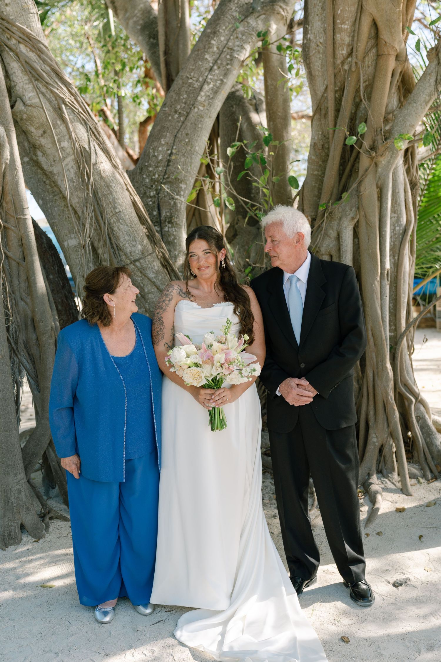 A bride and groom with an older woman, likely a parent, walking together in a tropical setting with large trees.