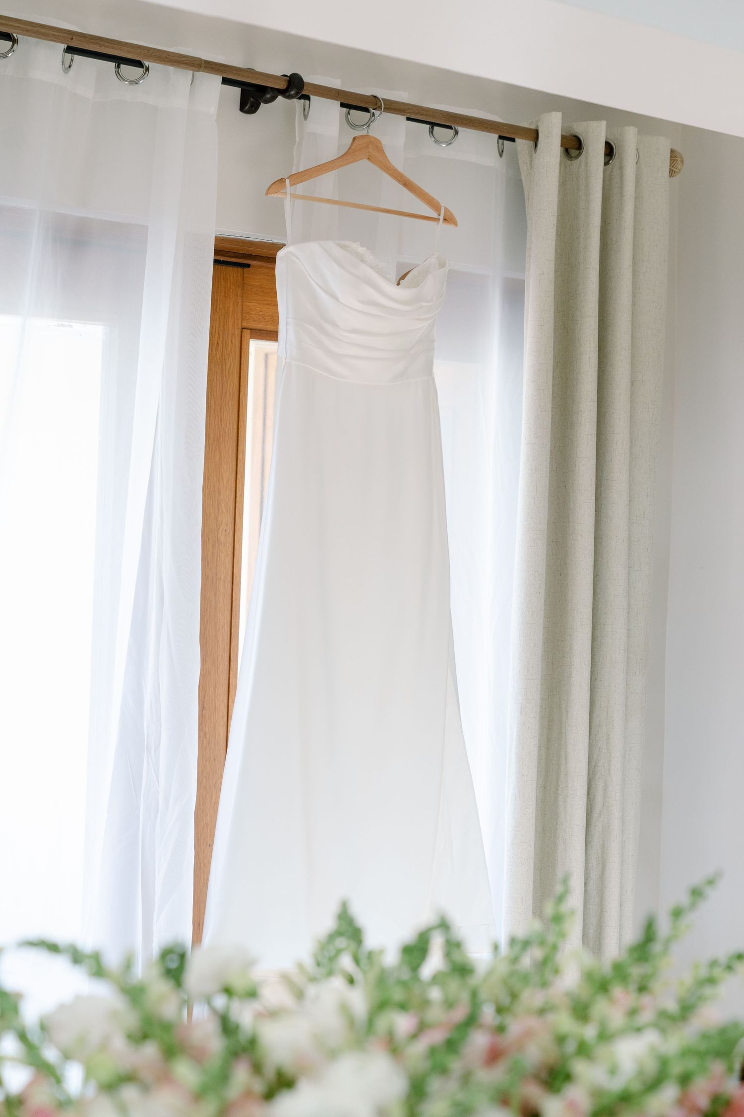 A white wedding dress hanging on a clothing rack in a room with potted plants.