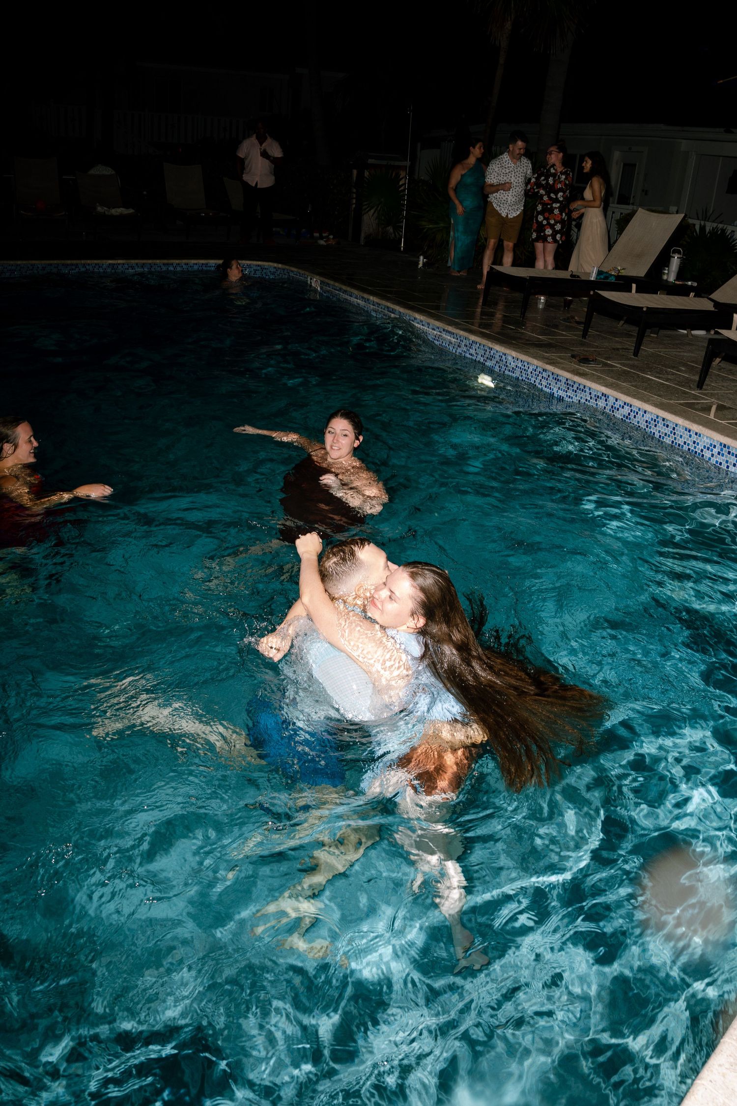Wedding guests enjoy a nighttime pool party with people swimming and splashing in illuminated turquoise water.
