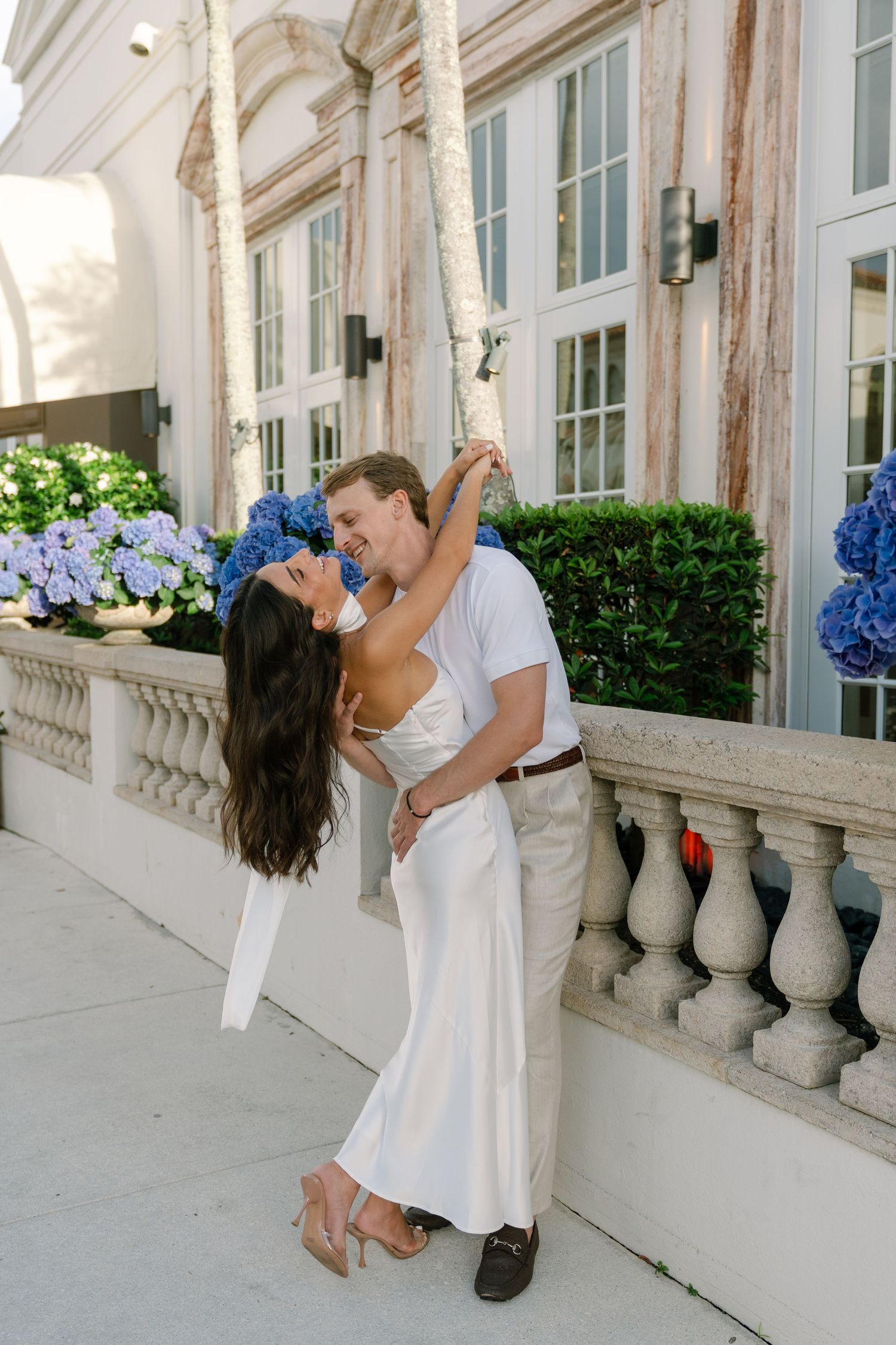 Couple sharing romantic kiss near elegant balustrade with blue hydrangeas, woman in white slip dress, man in white shirt.