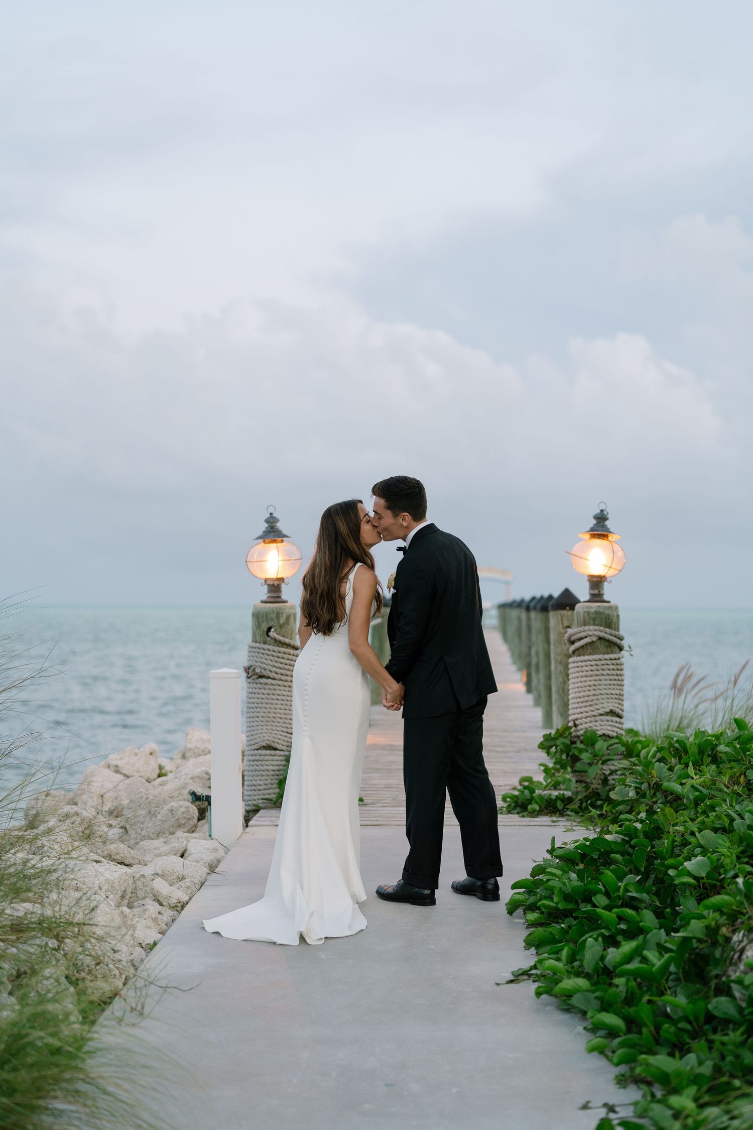 Bride and groom sharing a romantic kiss on a waterfront pier at dusk with glowing lanterns nearby.