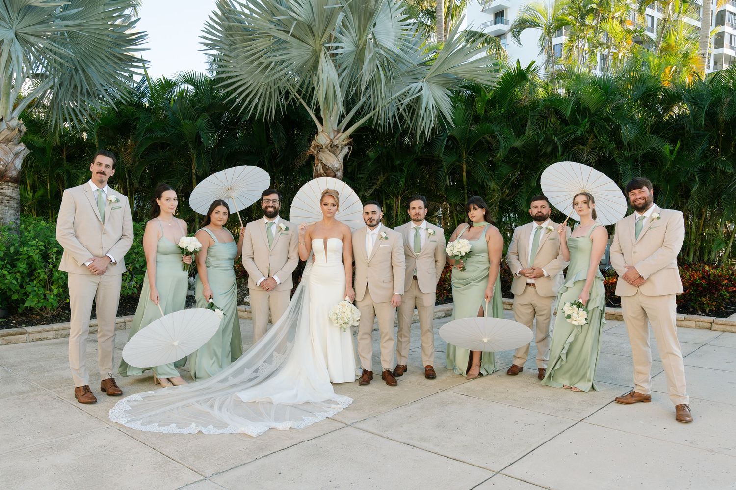 Wedding party in light colored attire poses outdoors with palm trees in the background.