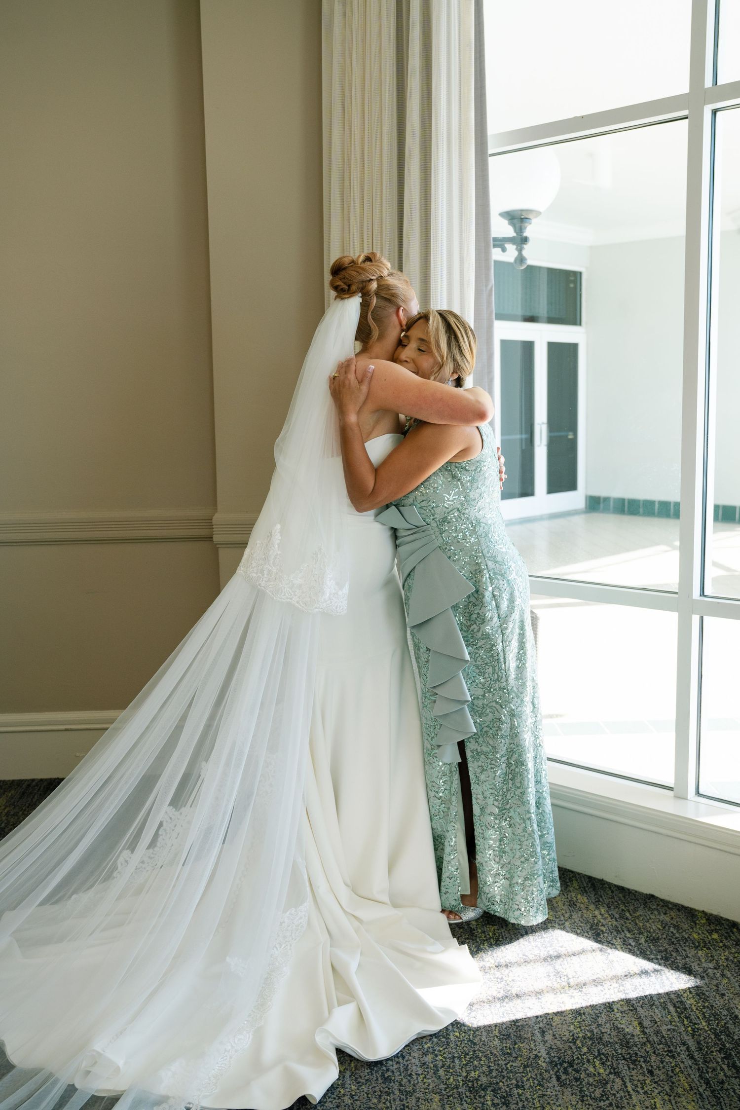 Two women share an emotional embrace in a sunlit room with a flowing white wedding veil.