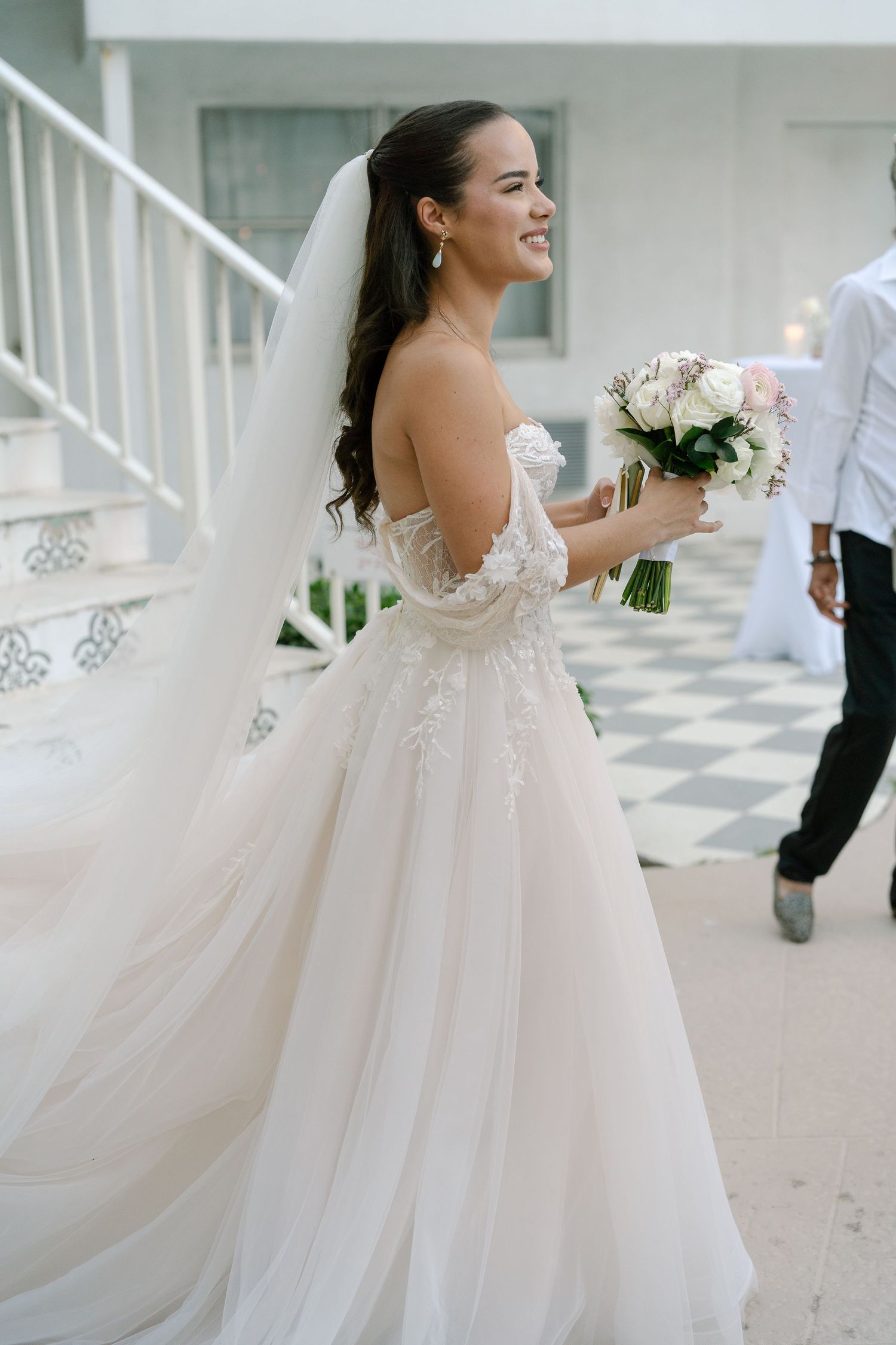 Smiling bride in a strapless lace gown holds a white bouquet on a checkered floor.