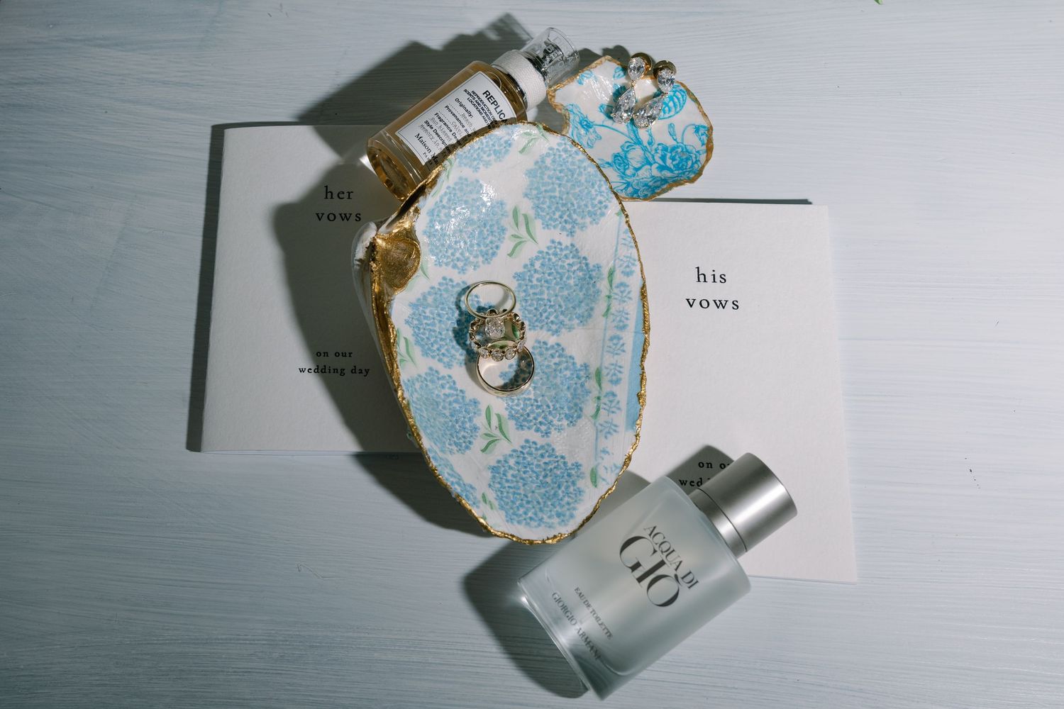 Wedding flat lay with floral ring box, his vows card, wedding rings, and Giorgio Armani cologne on white surface.