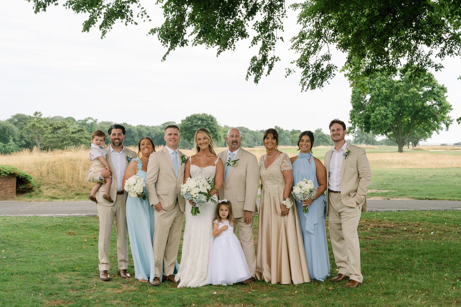 Wedding party poses outdoors in tan and blue attire with bride and groom centered on green lawn.