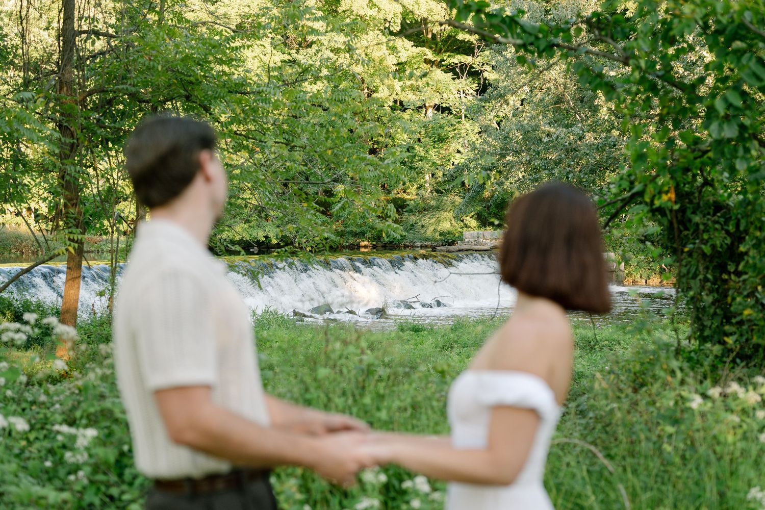 Couple holding hands near a serene waterfall surrounded by lush green trees outdoors.