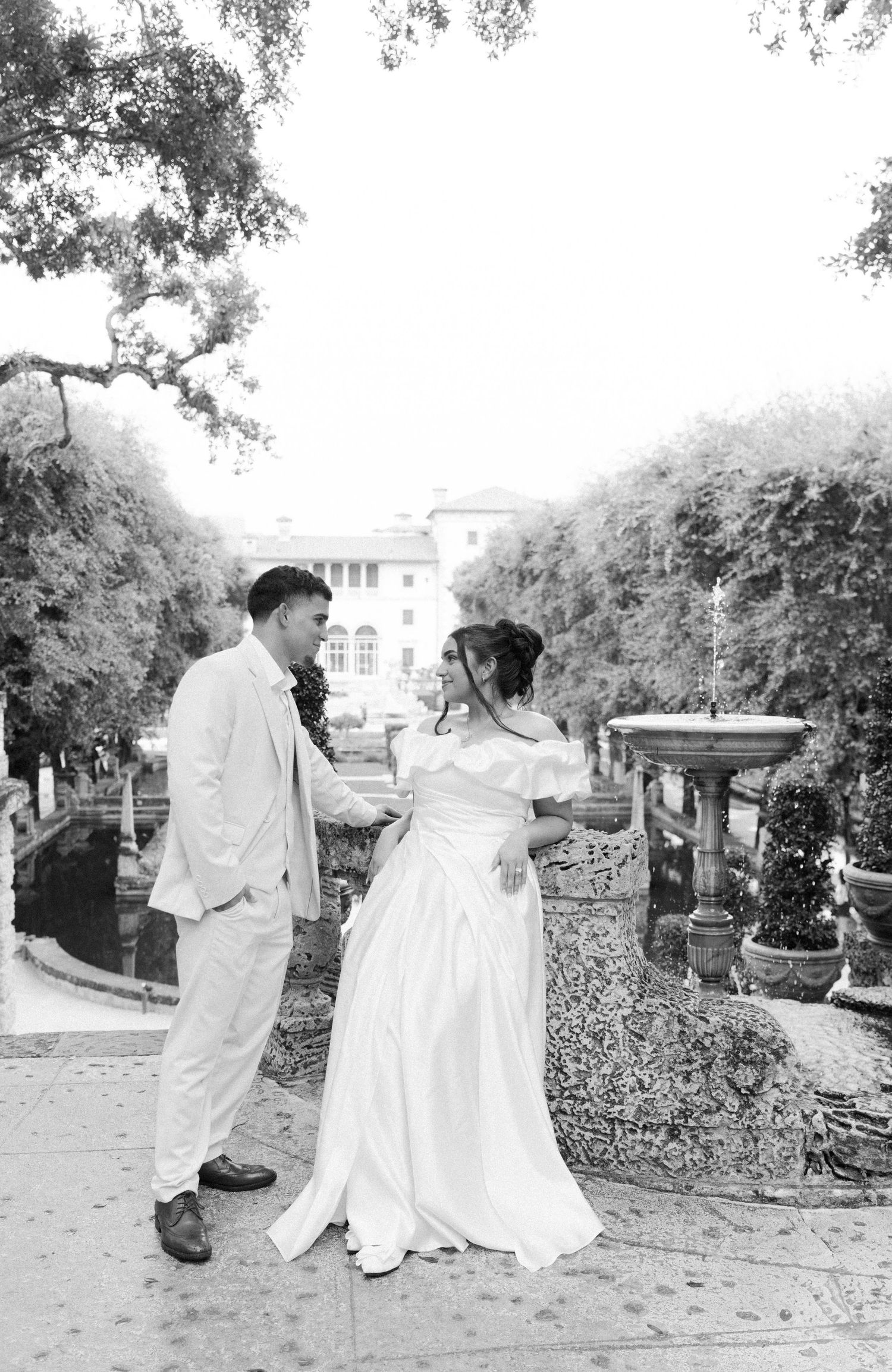 A black and white wedding photo showing a couple in formal attire standing outdoors in a garden setting.