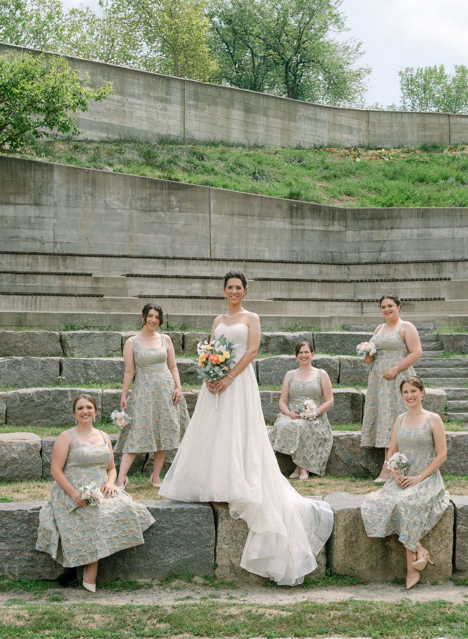 Bridal party poses on stone amphitheater steps in pale dresses with bouquets.