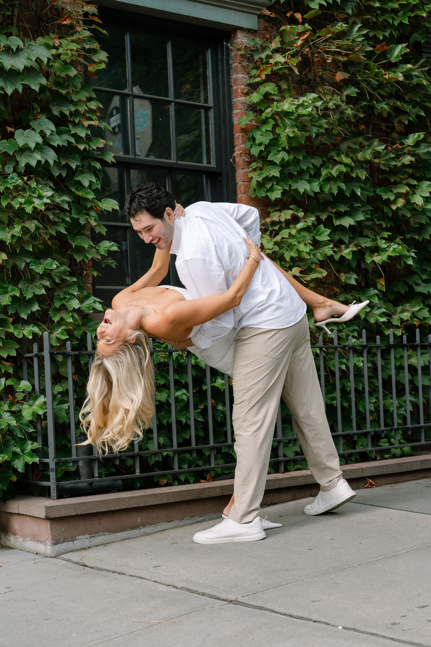 Couple dancing outdoors, man dipping woman with long blonde hair, ivy-covered brick wall background.