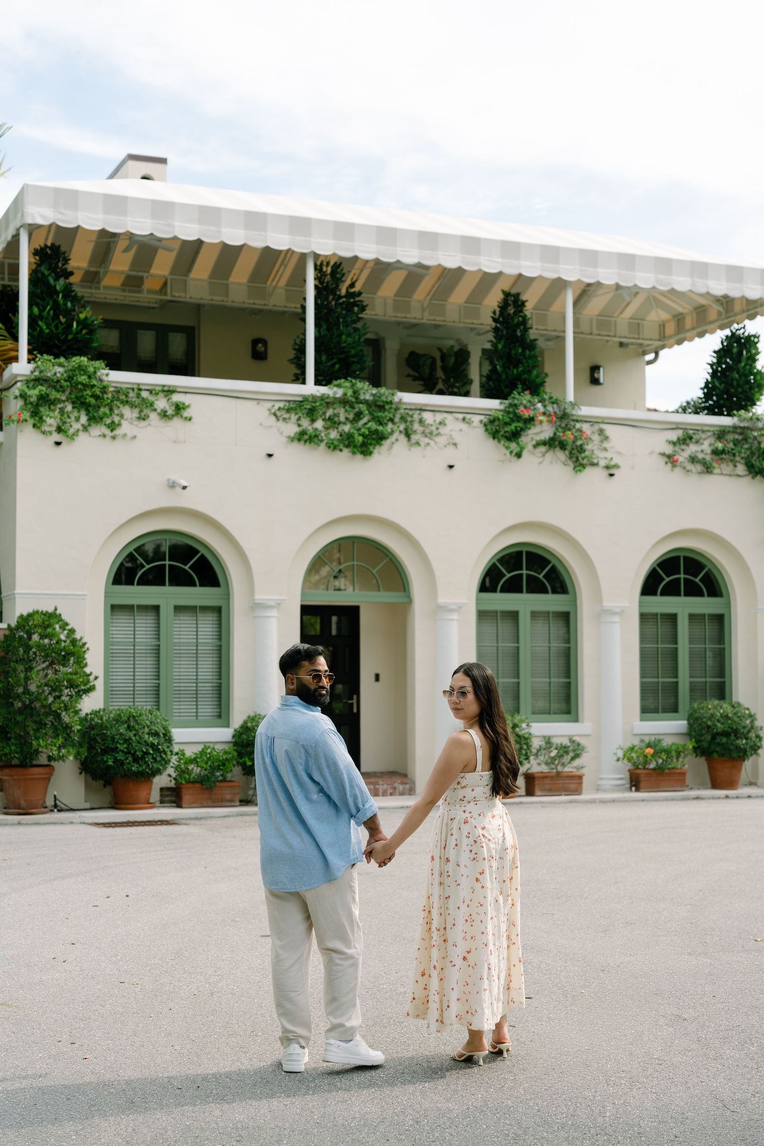 Couple holding hands in front of elegant white villa with arched windows and lush greenery, man in blue shirt and woman in floral dress.