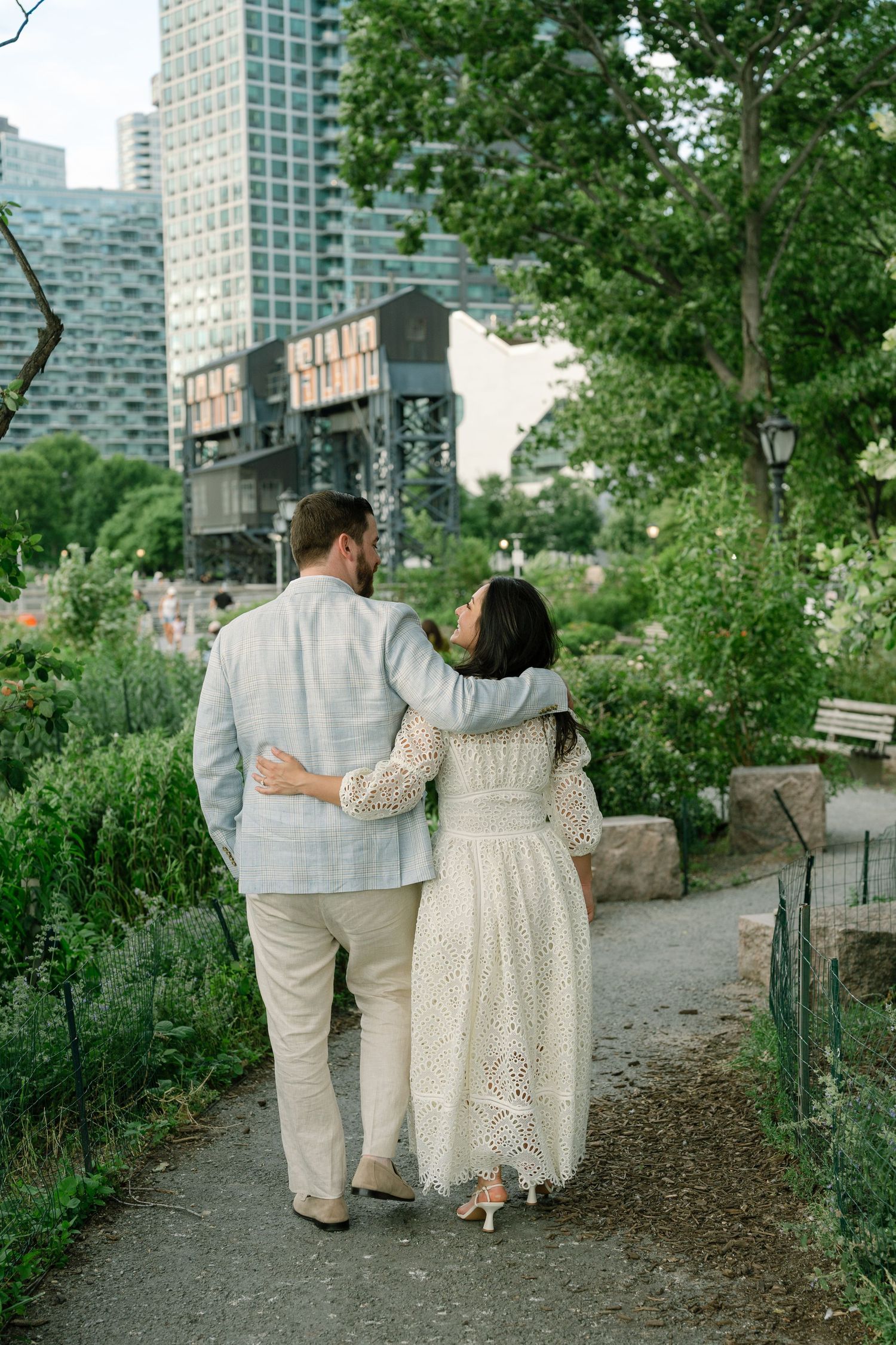 Couple in white outfits walking arm in arm through a lush urban park with city buildings in the background.