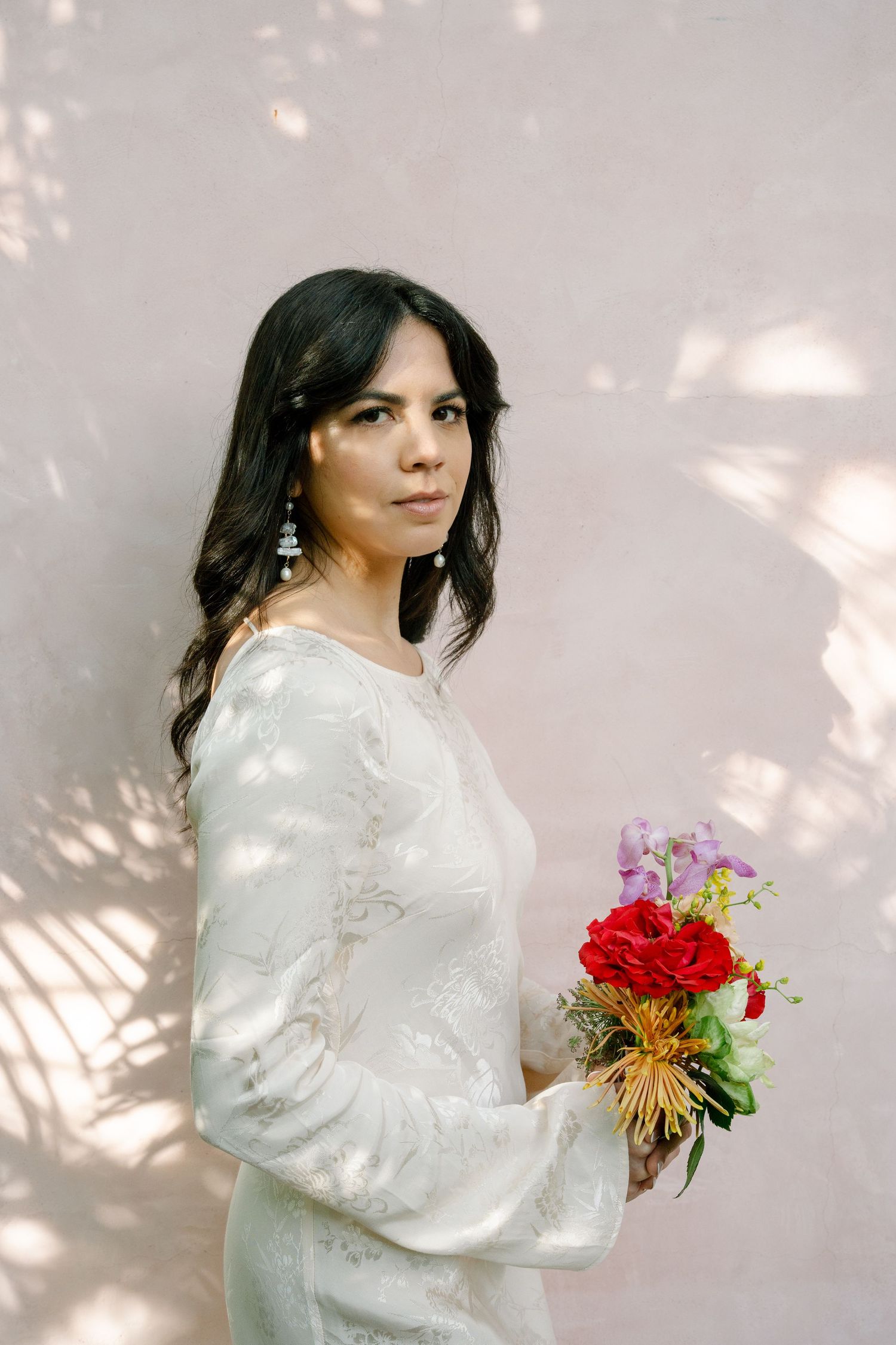 Elegant white long-sleeved dress with delicate floral bouquet against light wall with dappled shadows.