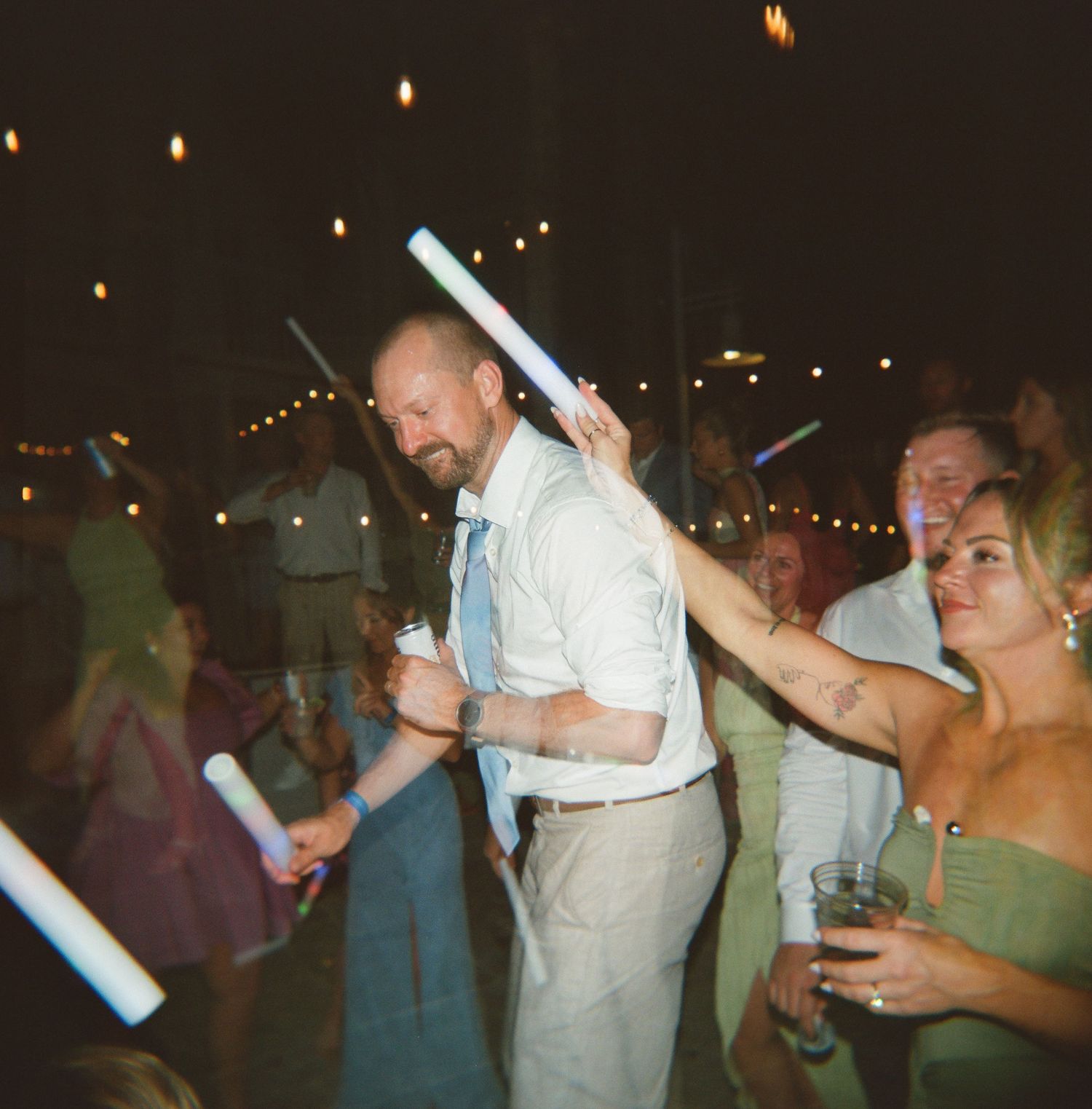 Party guests dance with glow sticks at a dimly lit wedding reception celebration.